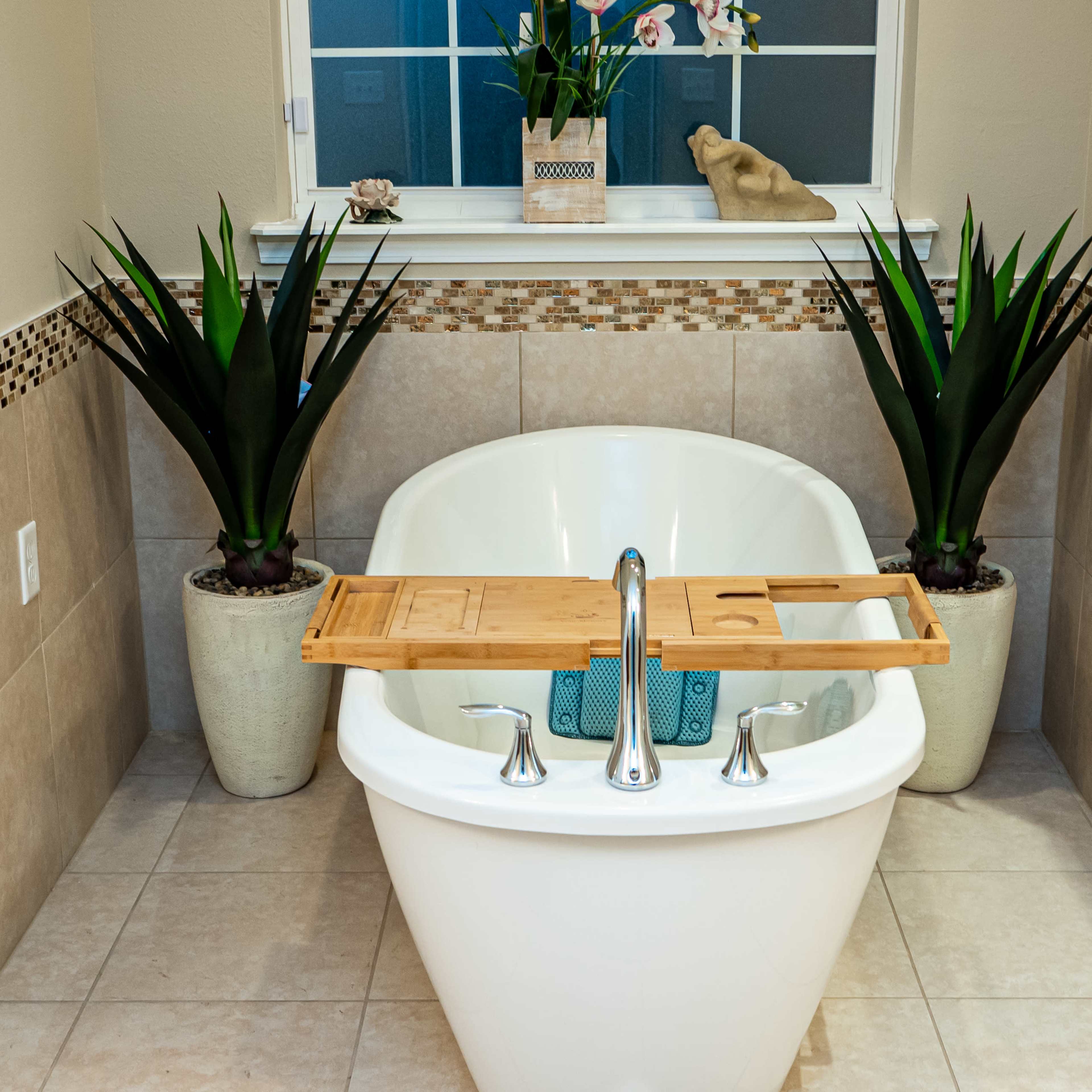 A freestanding white bathtub is centered in a neatly tiled bathroom, flanked by two tall plants and featuring a wooden tray across the top.