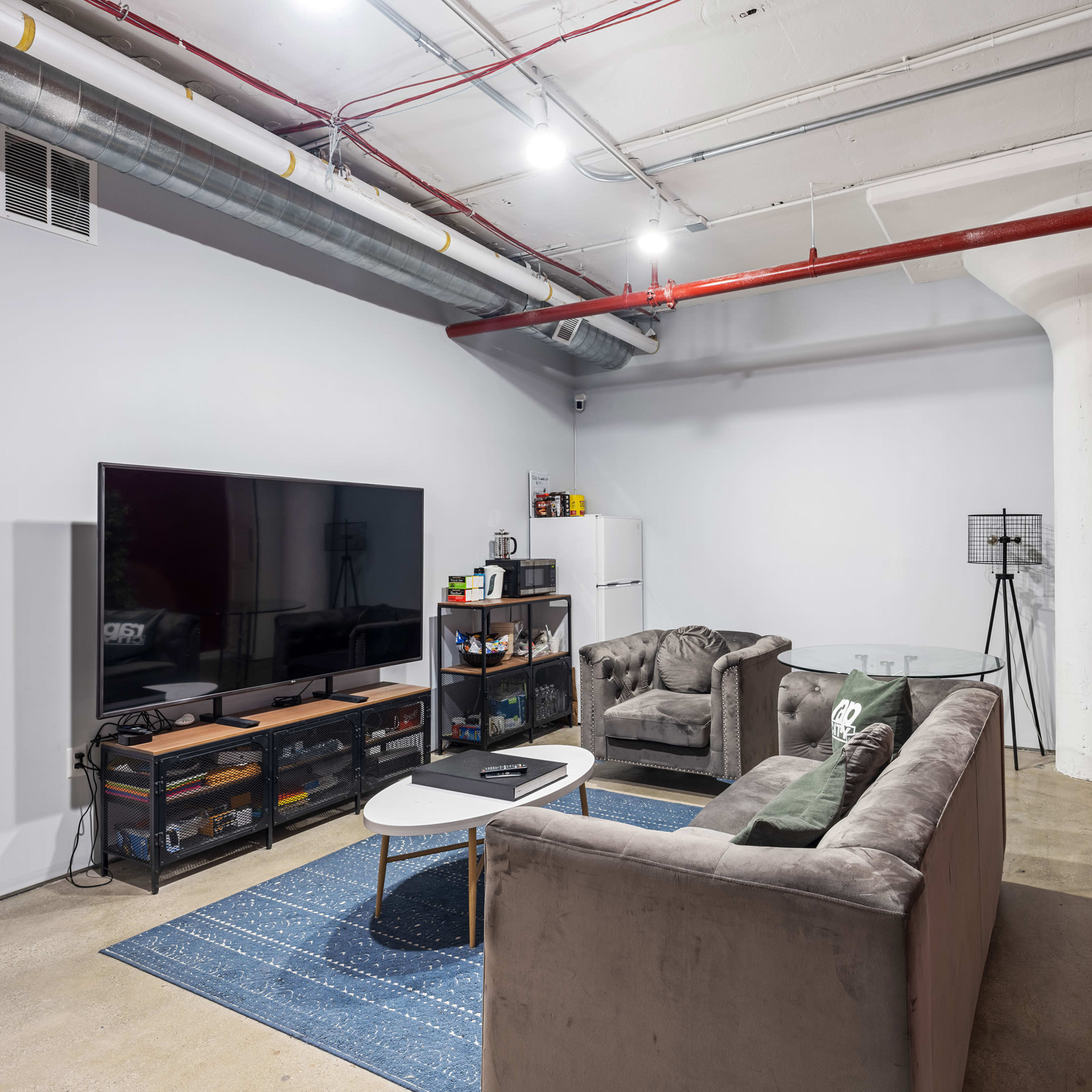 A spacious living room features a large television, two gray sofas, a coffee table, and a white refrigerator against one wall.