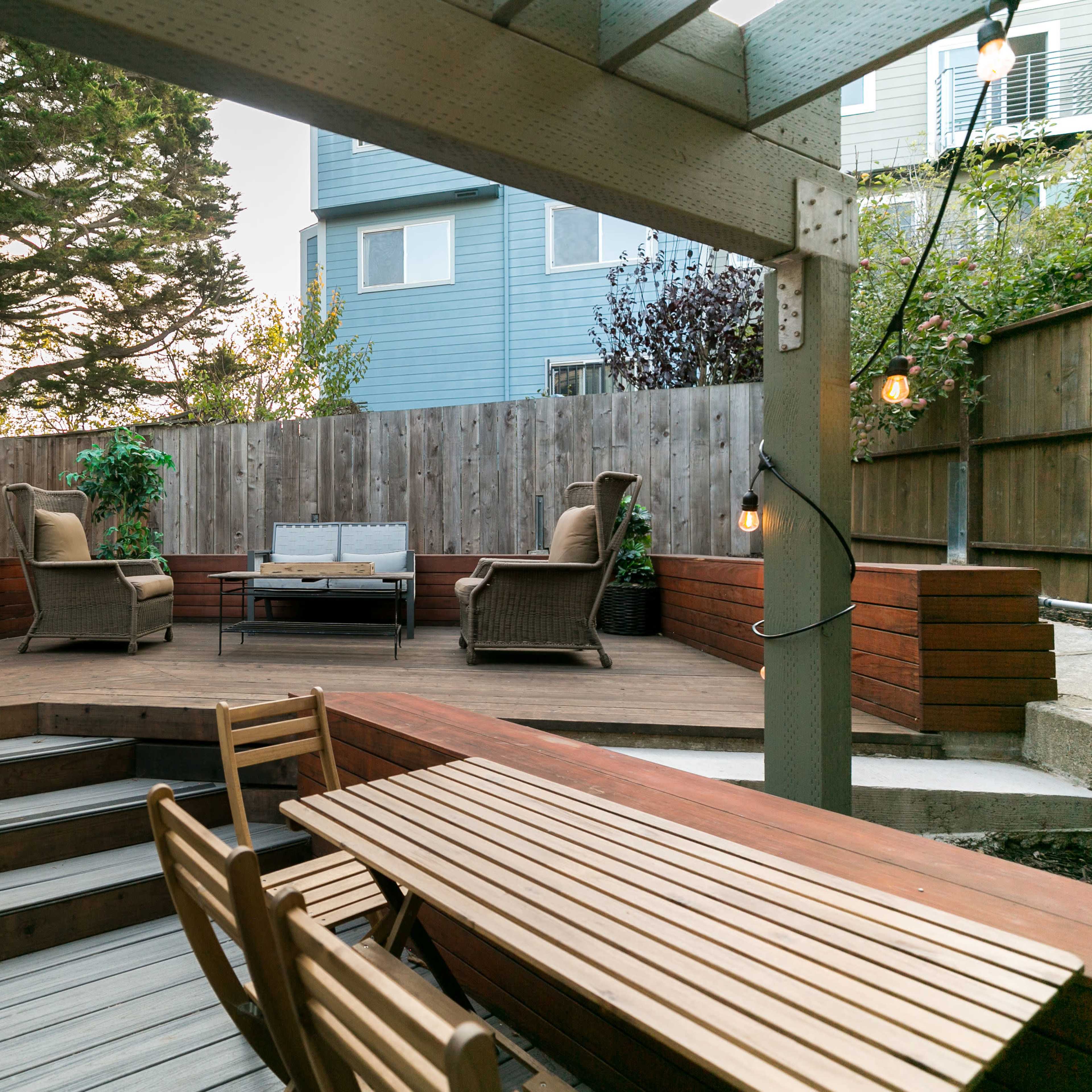 A wooden deck with a seating area, a coffee table, and string lights, surrounded by a wooden fence and residential buildings.