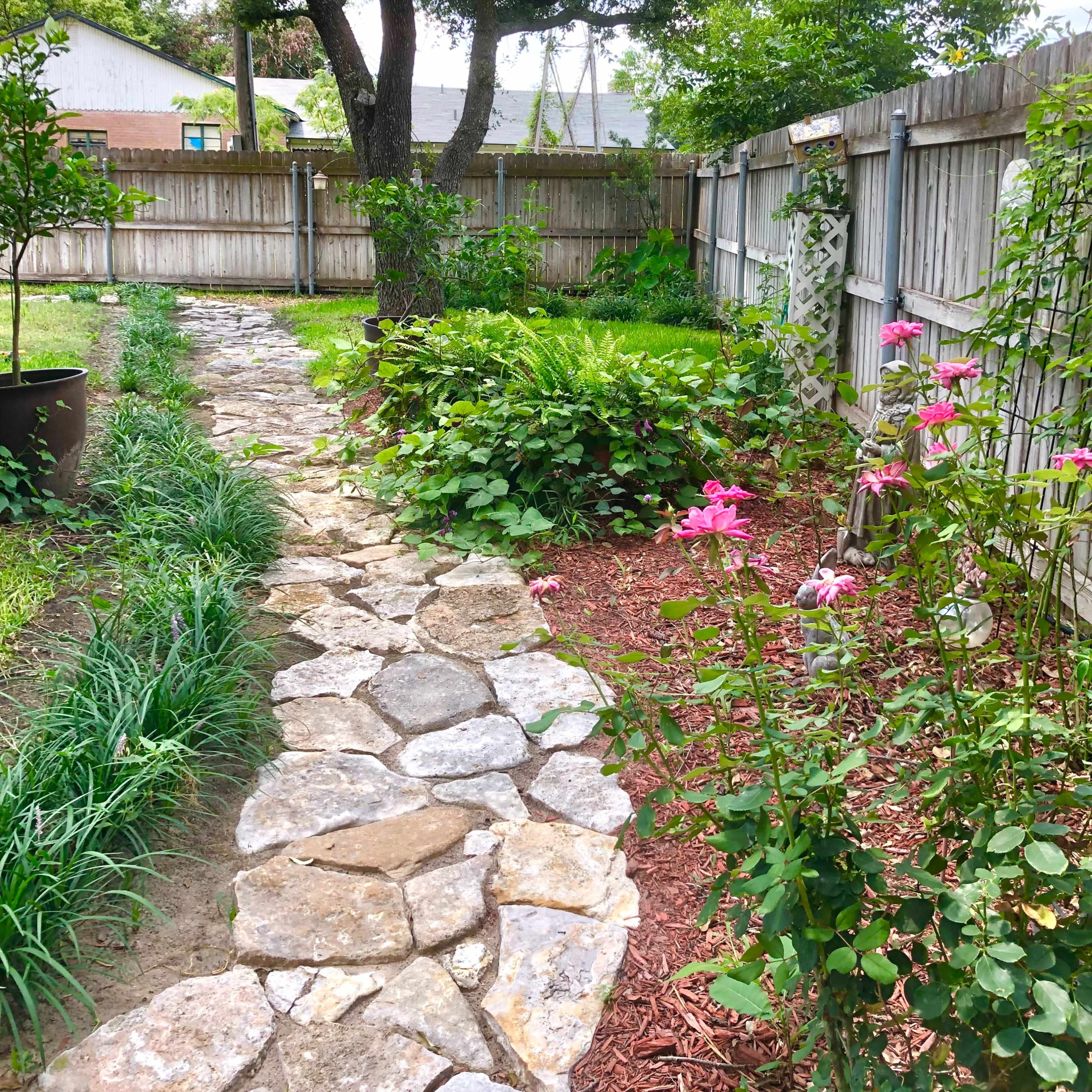 A stone pathway weaves through a lush garden bordered by a wooden fence, with flowering plants and green foliage lining the sides.