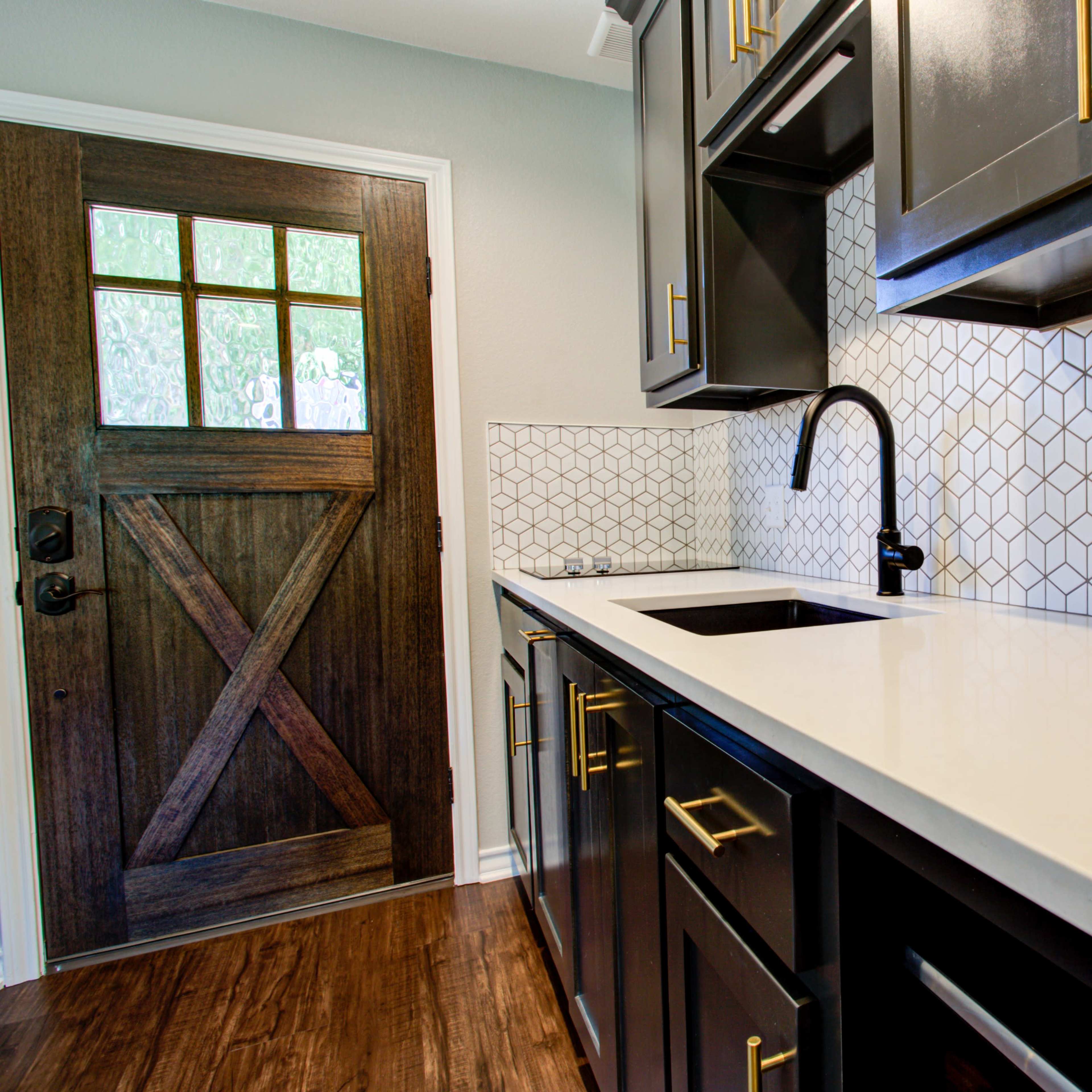 A kitchen features dark cabinets, a white countertop, a geometric backsplash, and a coffee maker next to a wooden door.
