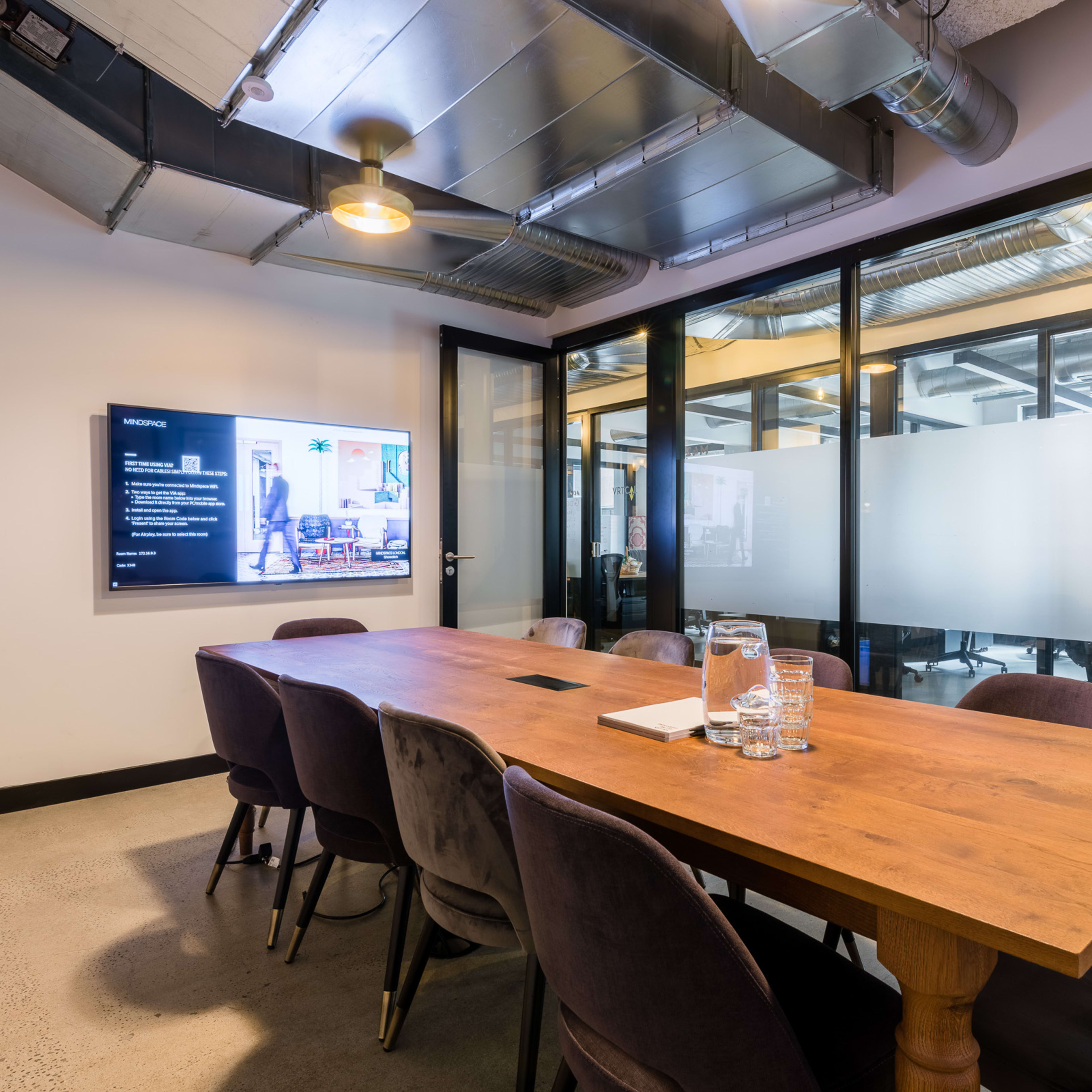 A modern meeting room with a wooden table surrounded by upholstered chairs, a wall-mounted screen, and glass partitions.