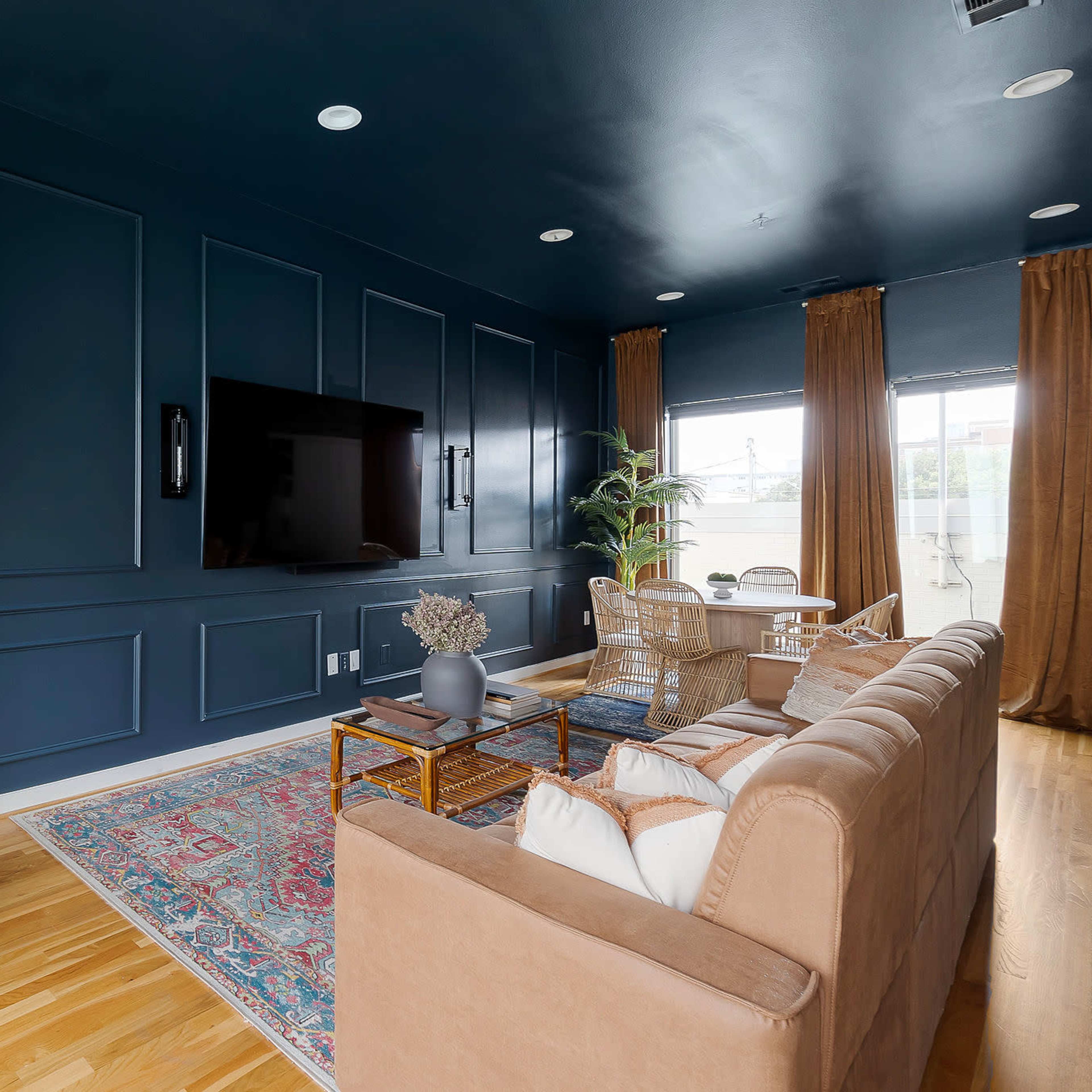 A modern living room featuring dark blue paneled walls, a sofa and armchair, a wooden coffee table, and large windows with curtains.