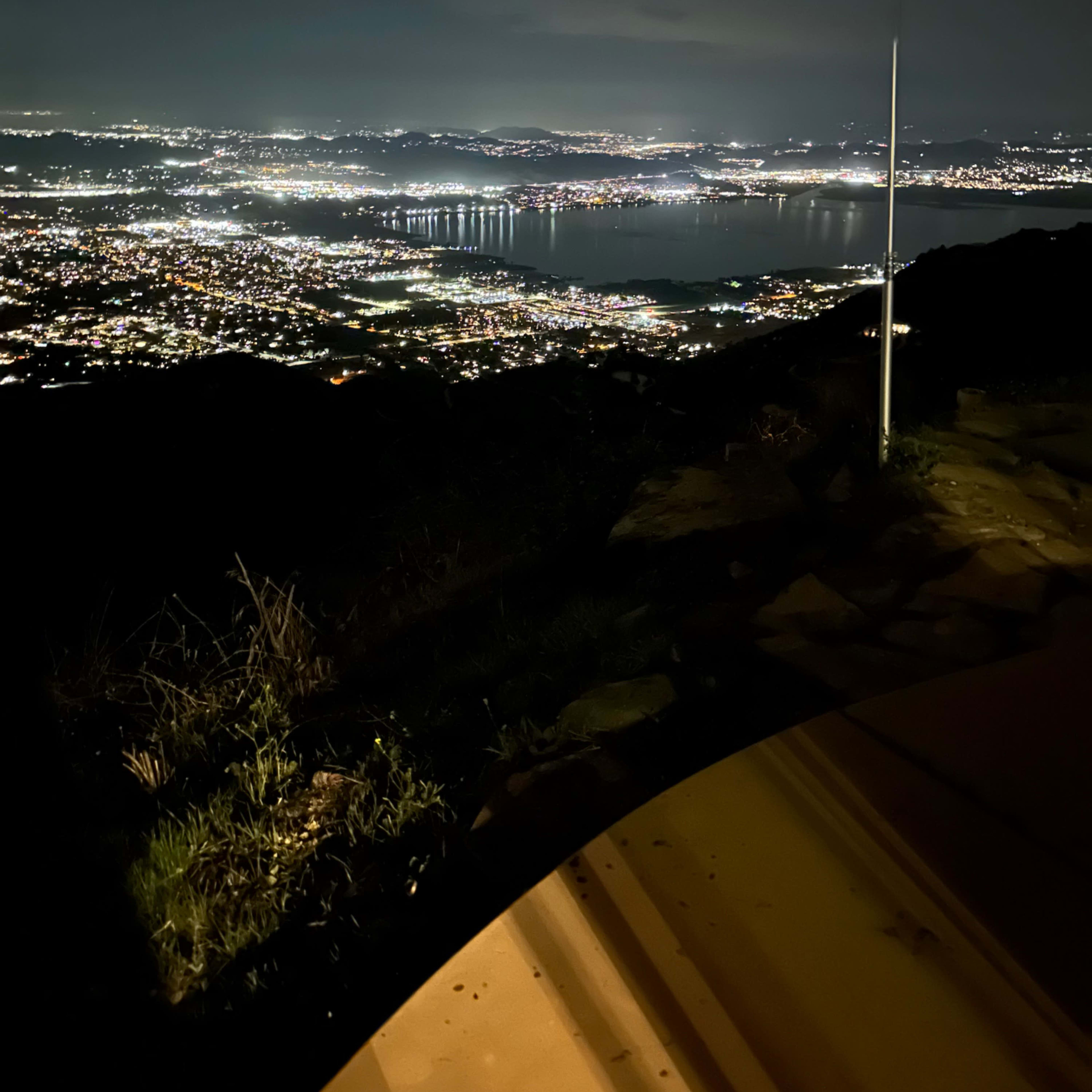 A nighttime panoramic view of a city landscape illuminated by lights, with a body of water and mountains visible in the distance.