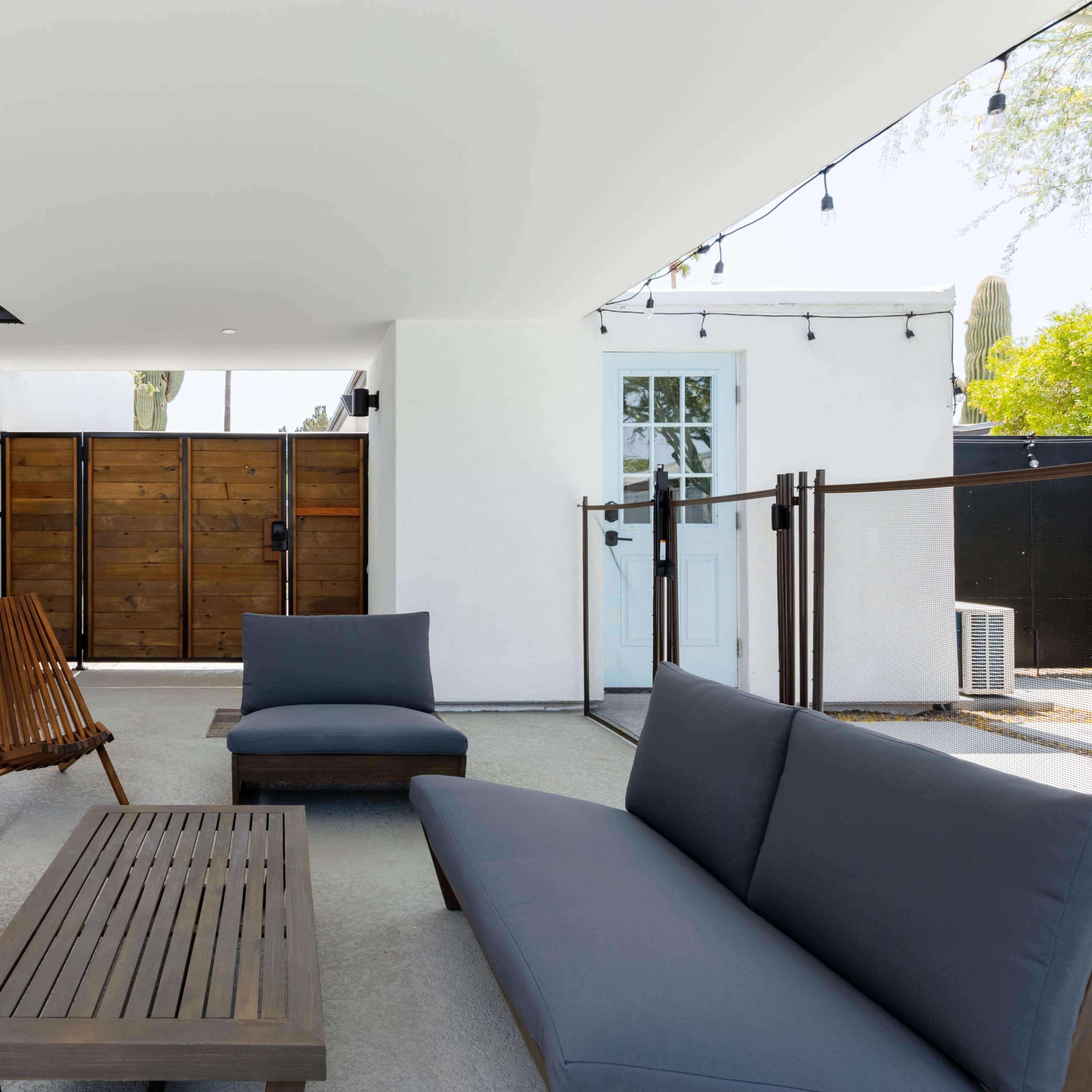 A modern outdoor seating area featuring two gray sofas, a wooden coffee table, and a wooden chair, all situated under a covered patio with a view of a wooden gate.