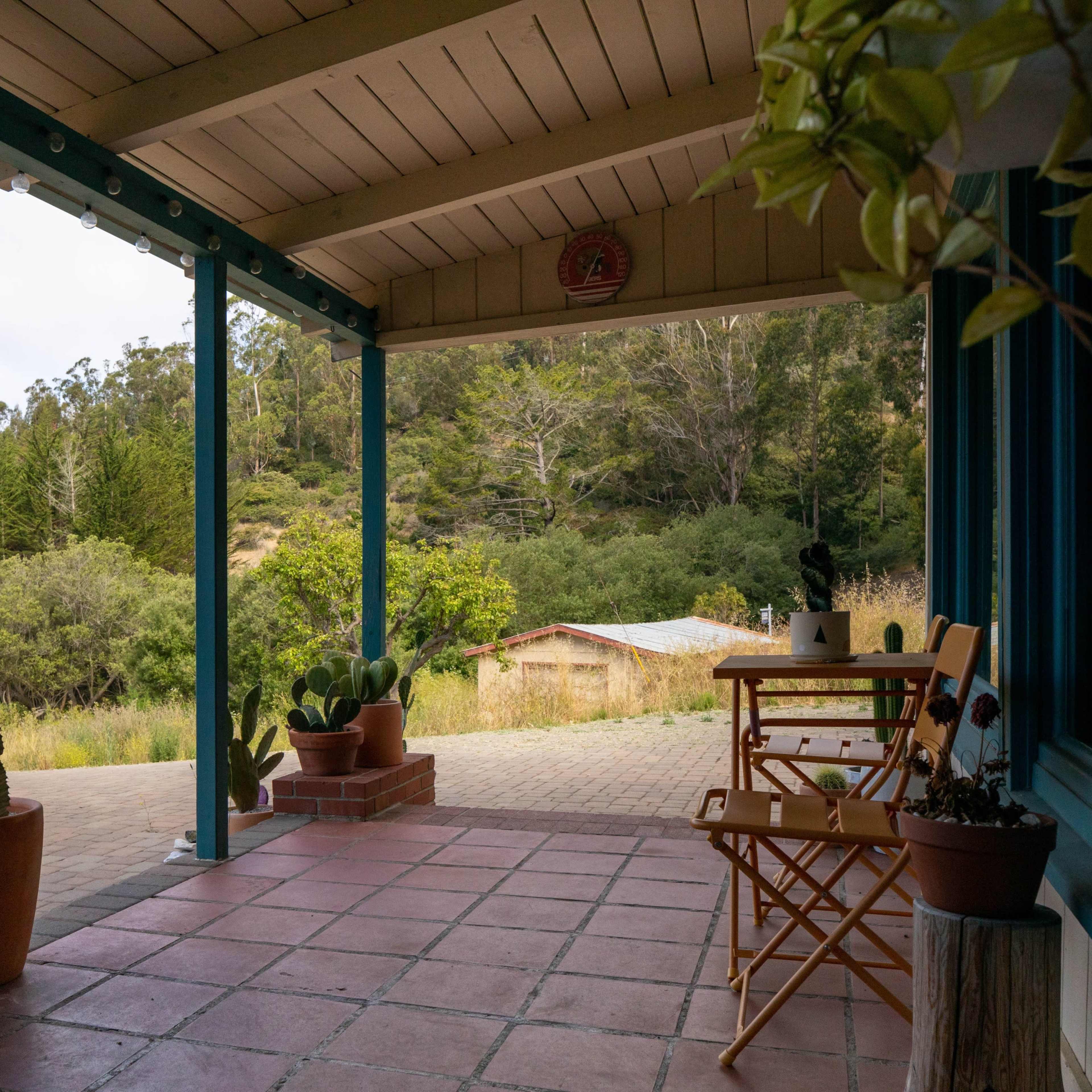 A porch with blue trim features potted cacti and a small wooden table and chair, overlooking a natural landscape.