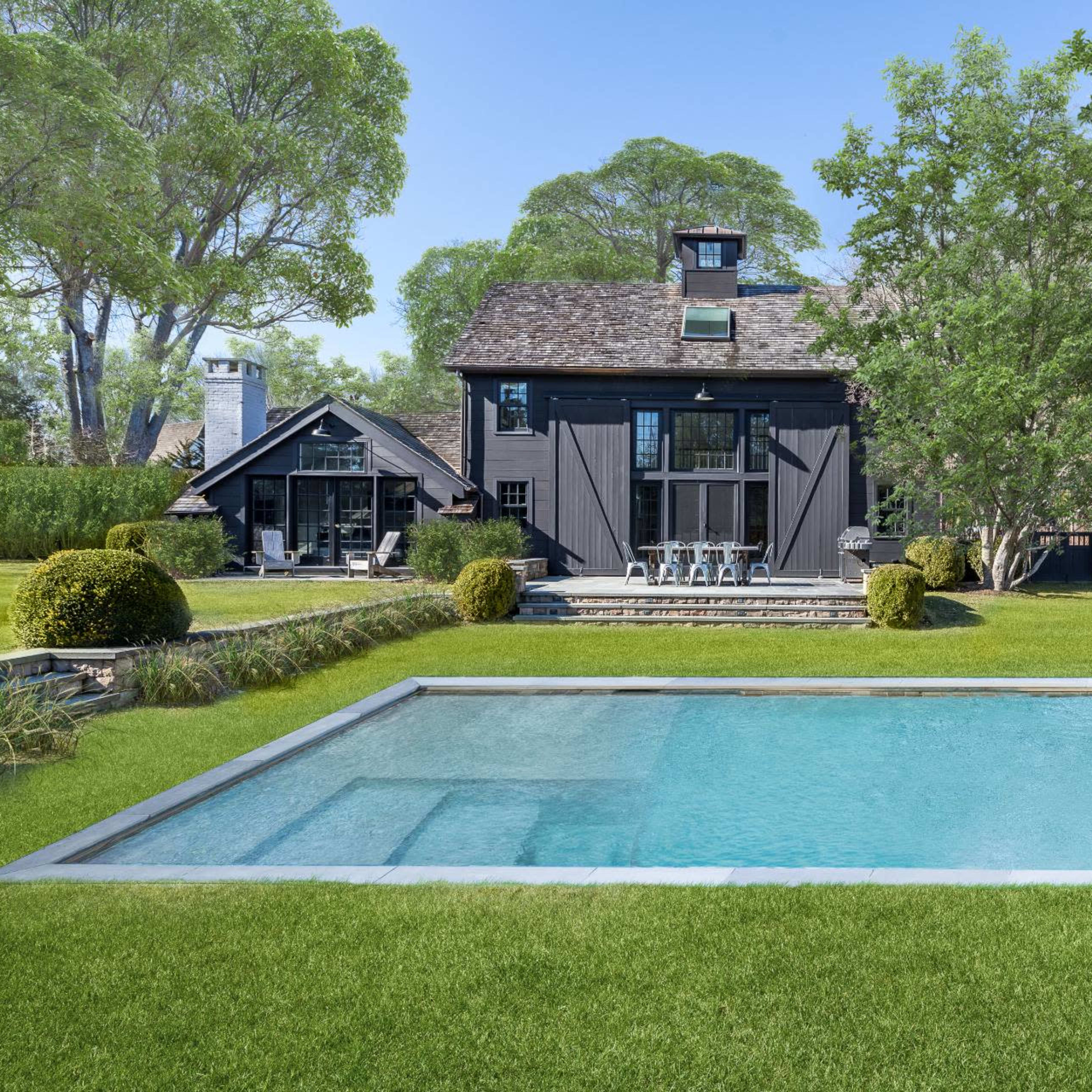 A modern black house with a pool is surrounded by lush green grass and trees.