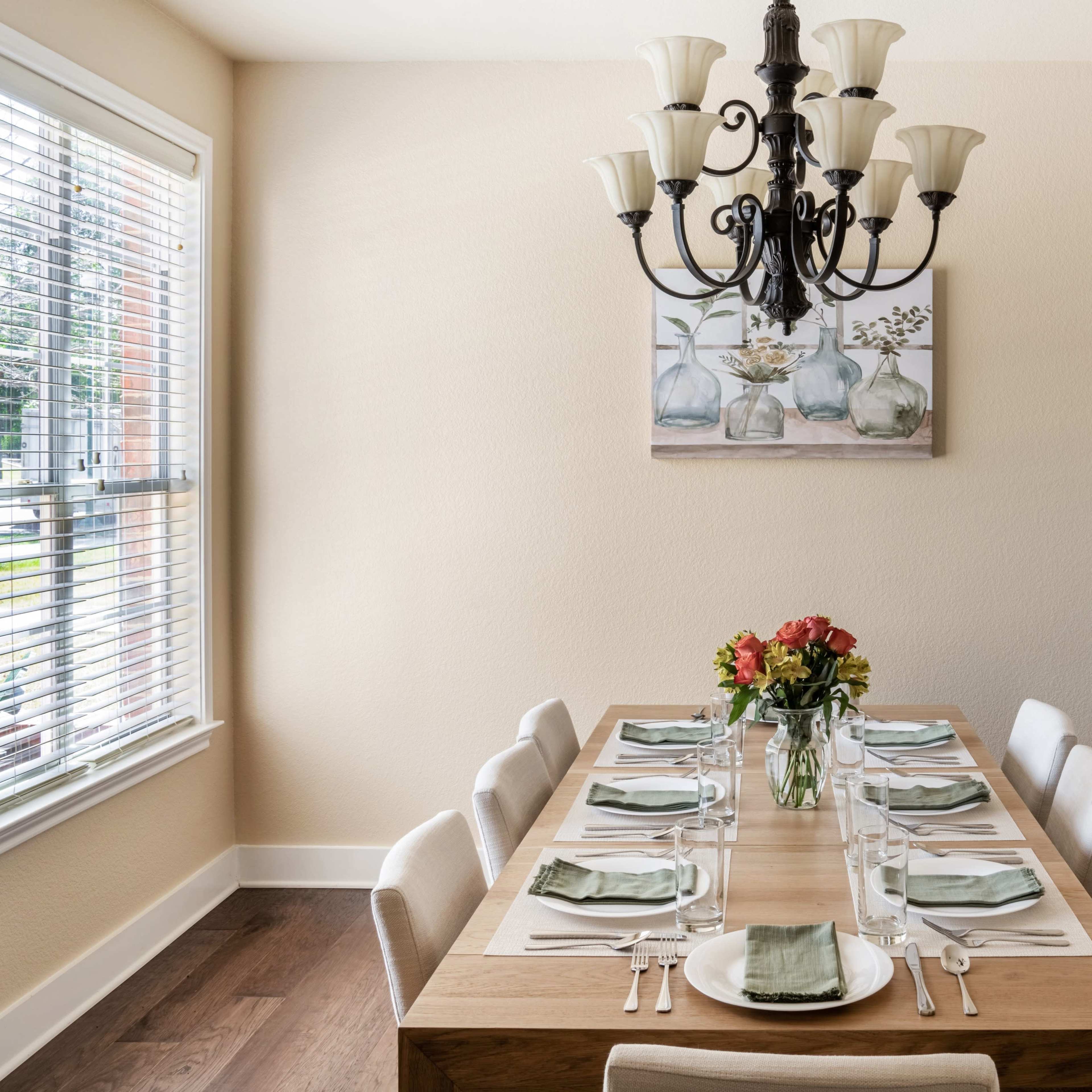 A dining room features a large wooden table set for eight, with a chandelier overhead and a floral painting on the wall.