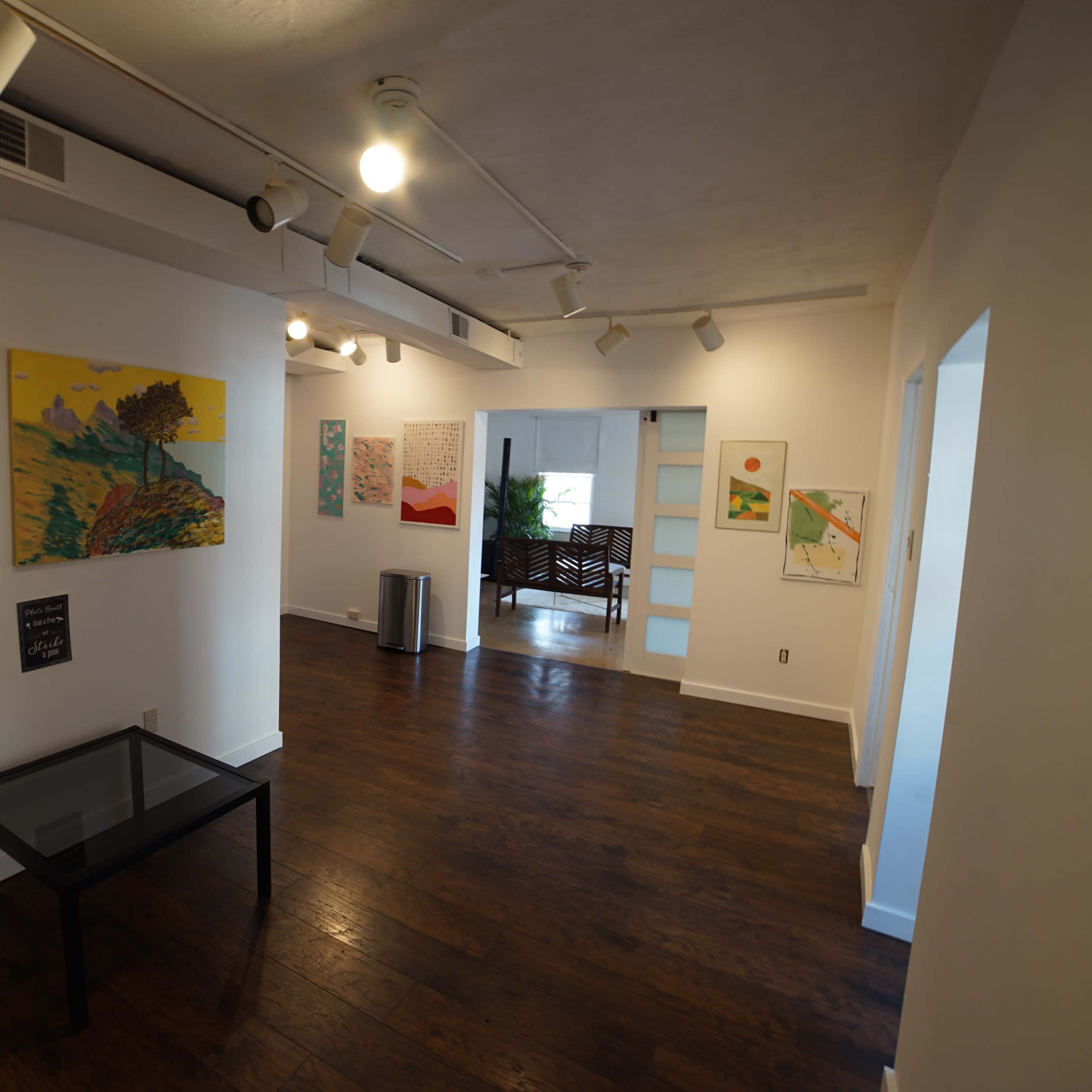 A well-lit gallery with several paintings on the white walls and a glass table in the foreground.