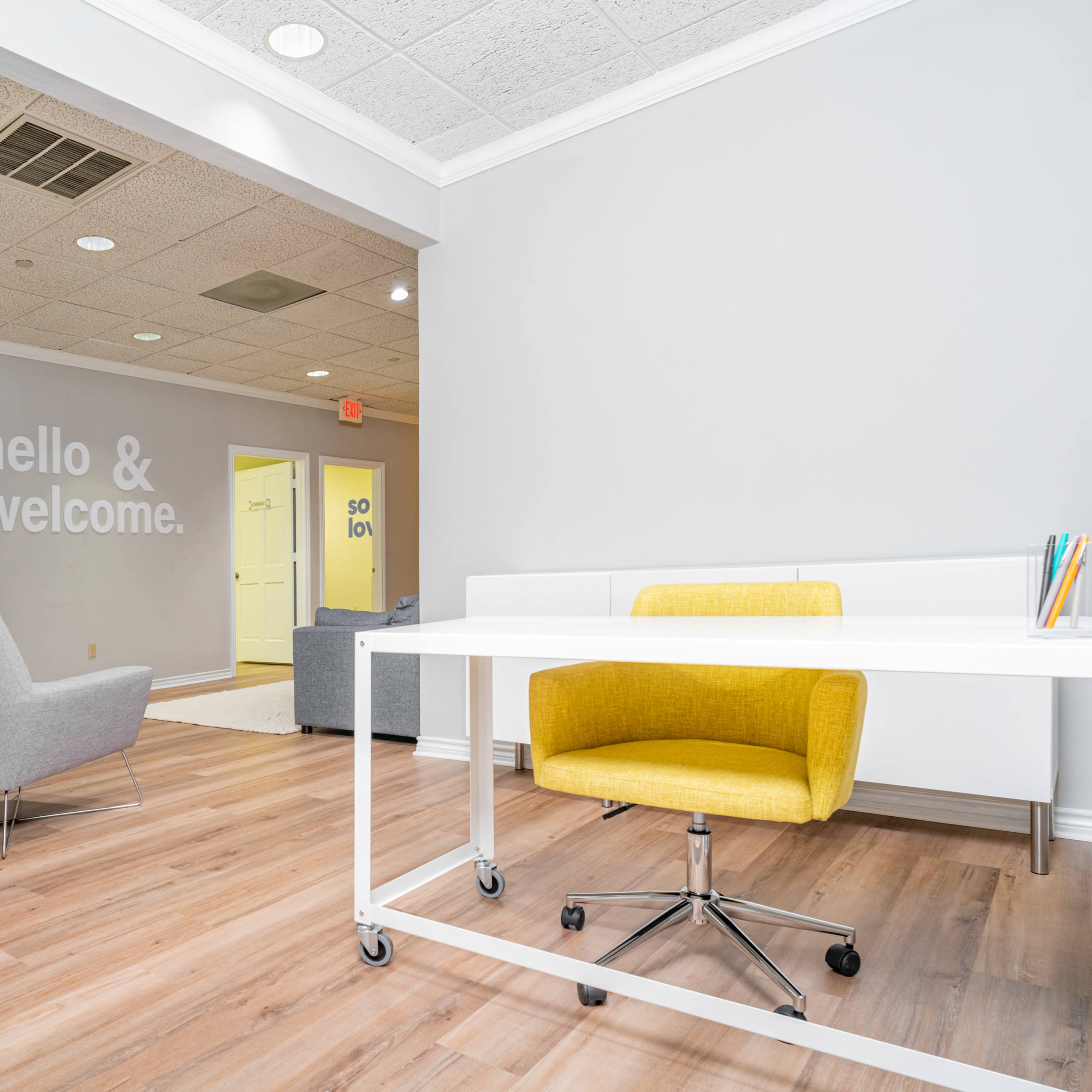 A modern office reception area with a white desk and a yellow chair, leading into a hallway with light-colored walls and welcoming signage.