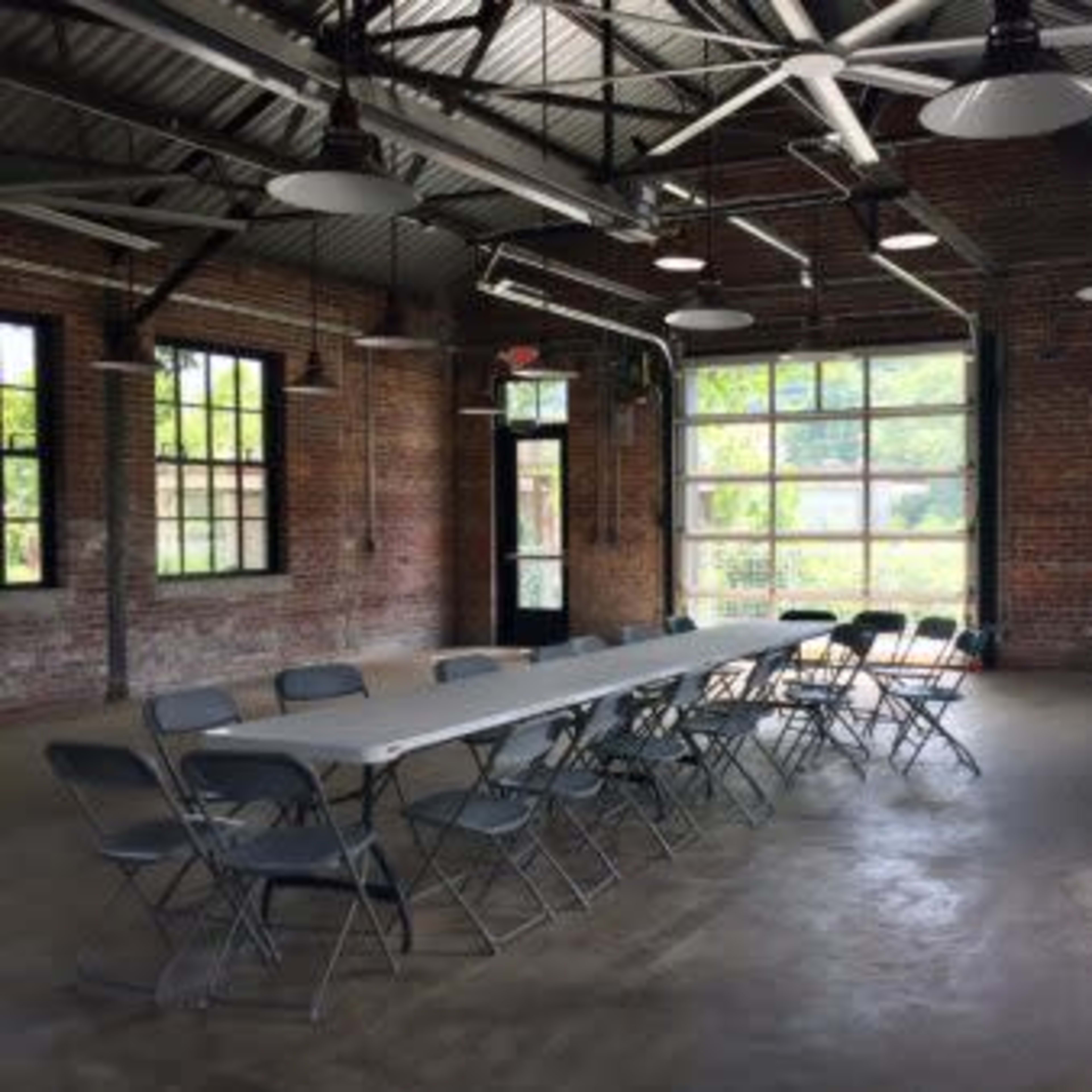 A long table with folding chairs is set up in an industrial-style room featuring exposed brick walls and large windows.