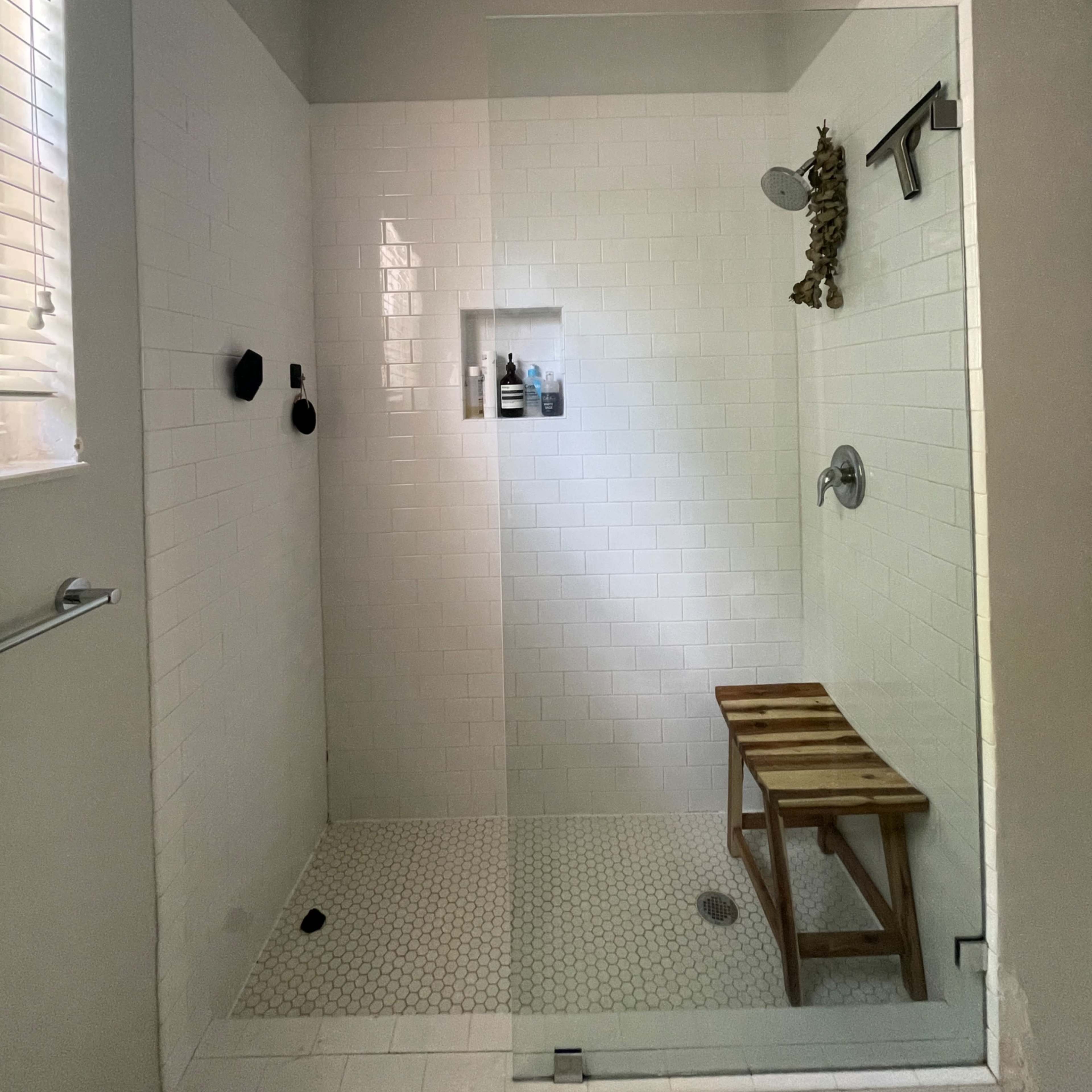 A modern shower with white tiled walls, a glass door, a wooden stool, and a shower head.