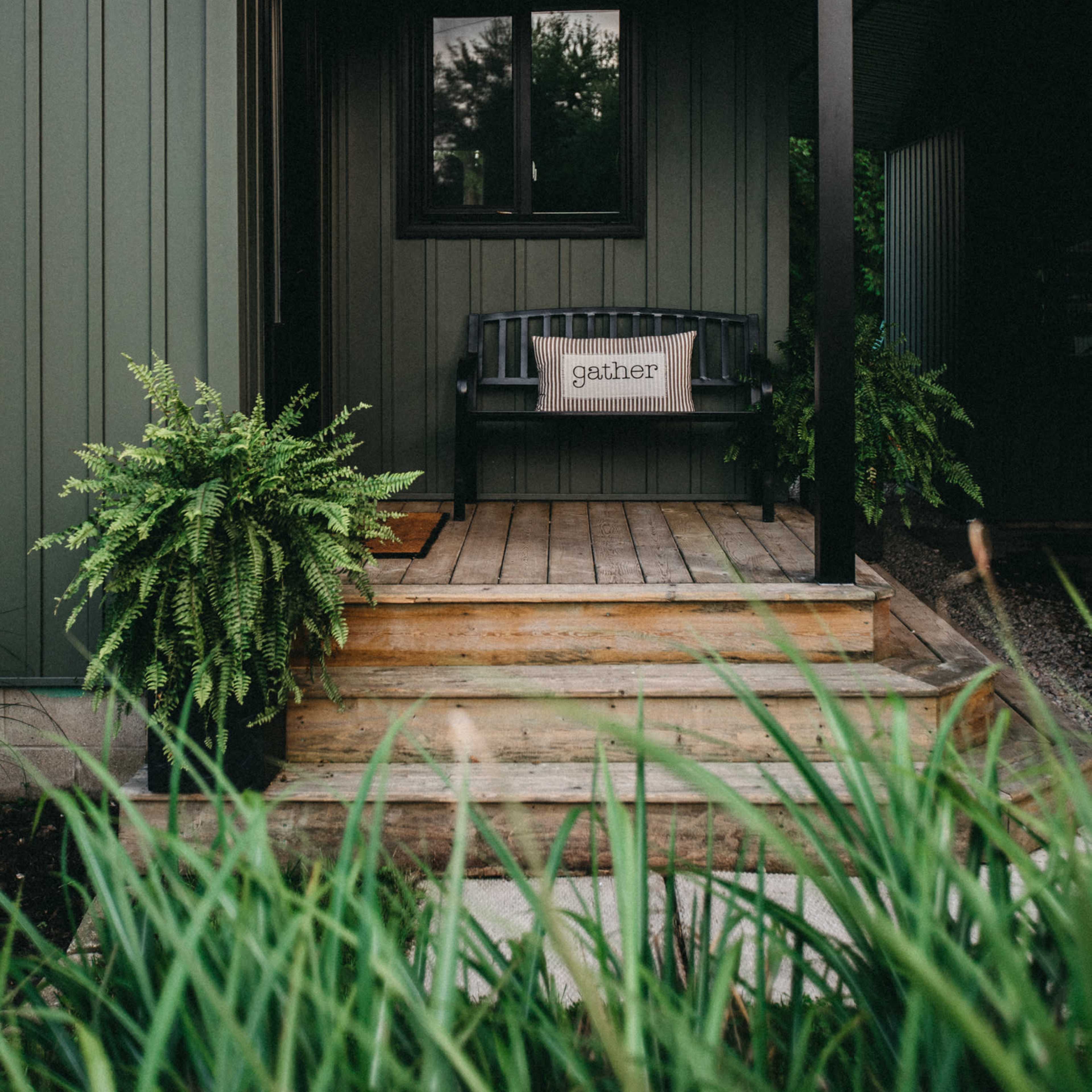A dark green house features a front porch with a wooden bench and a decorative sign that says "gather."