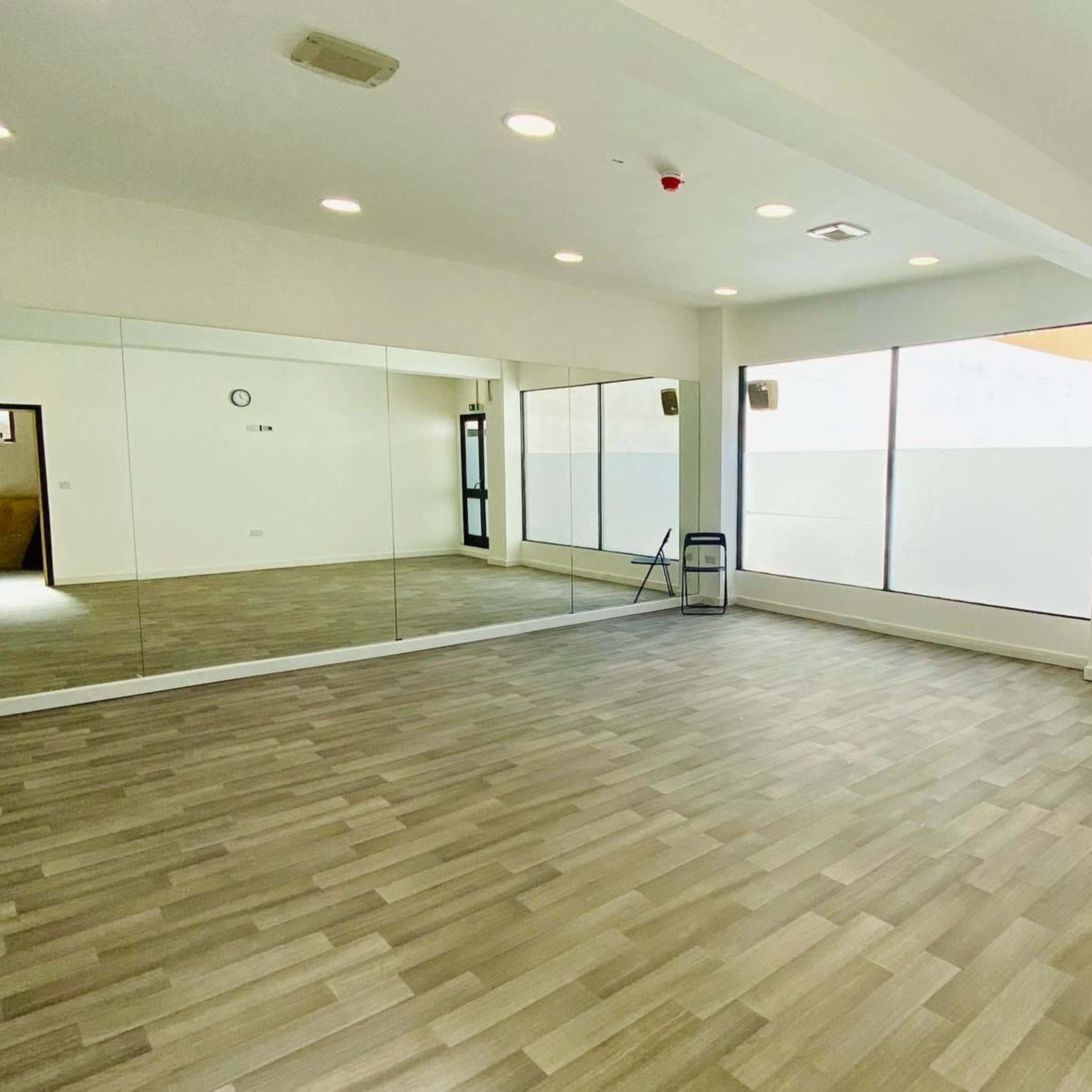 An empty room with wooden flooring, large windows, and a wall of mirrors.