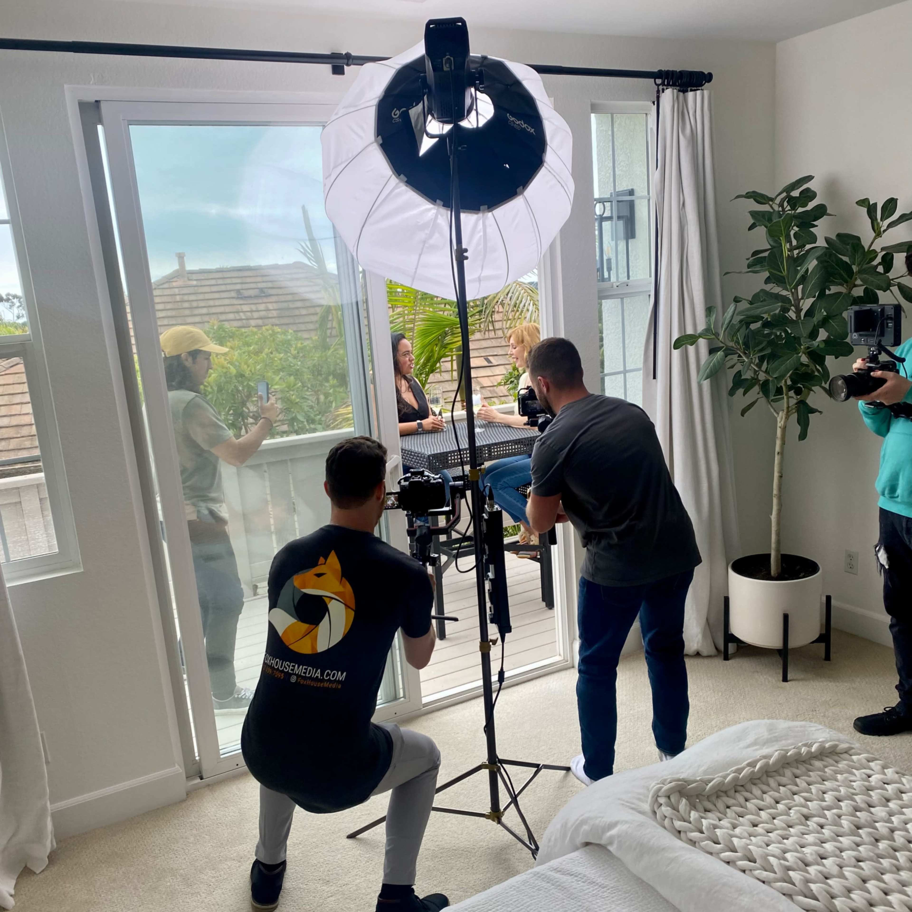 A film crew sets up equipment in a well-lit bedroom to shoot a scene featuring a person outside on a balcony.