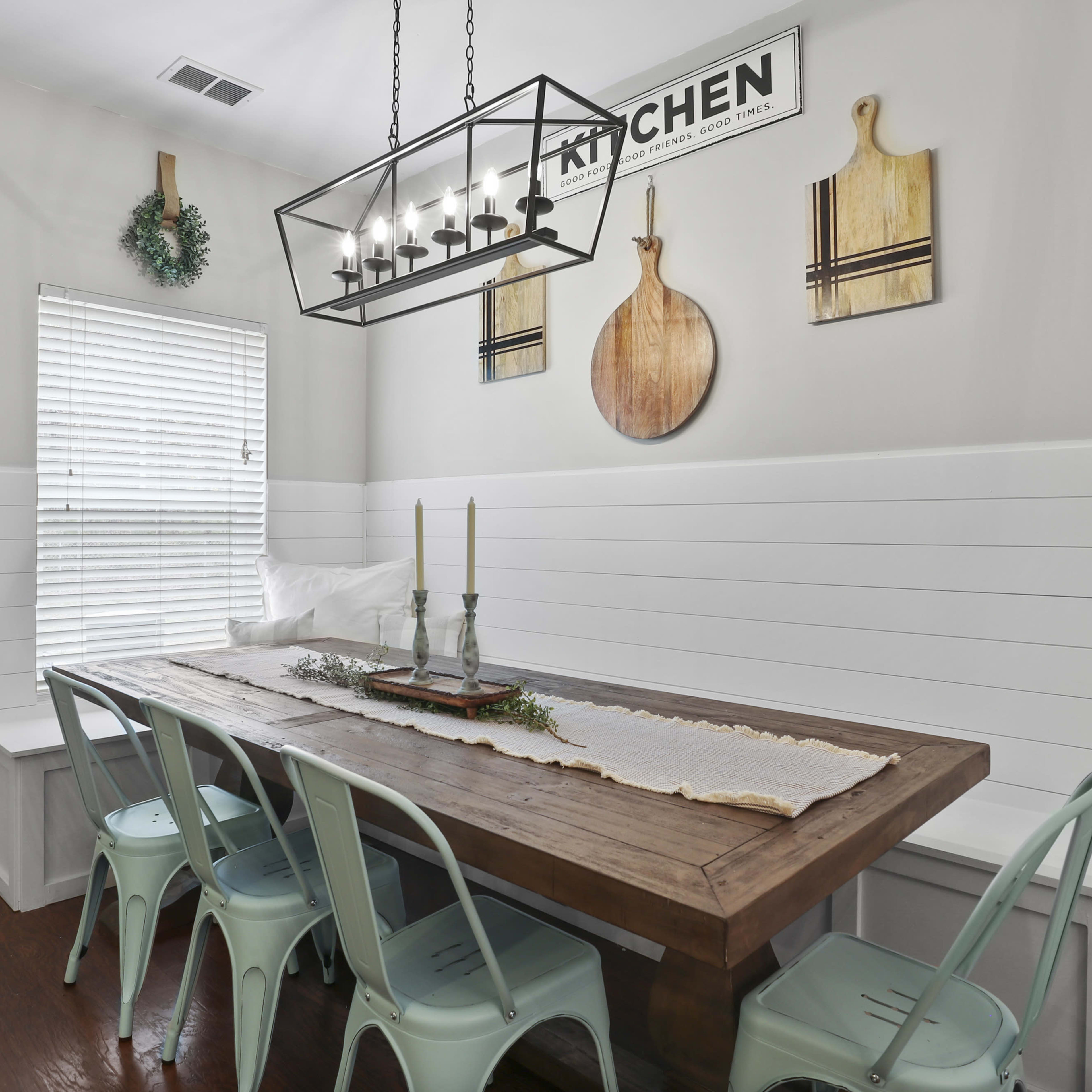 A dining area features a wooden table surrounded by light-colored chairs, with a chandelier above and a window adorned with blinds.
