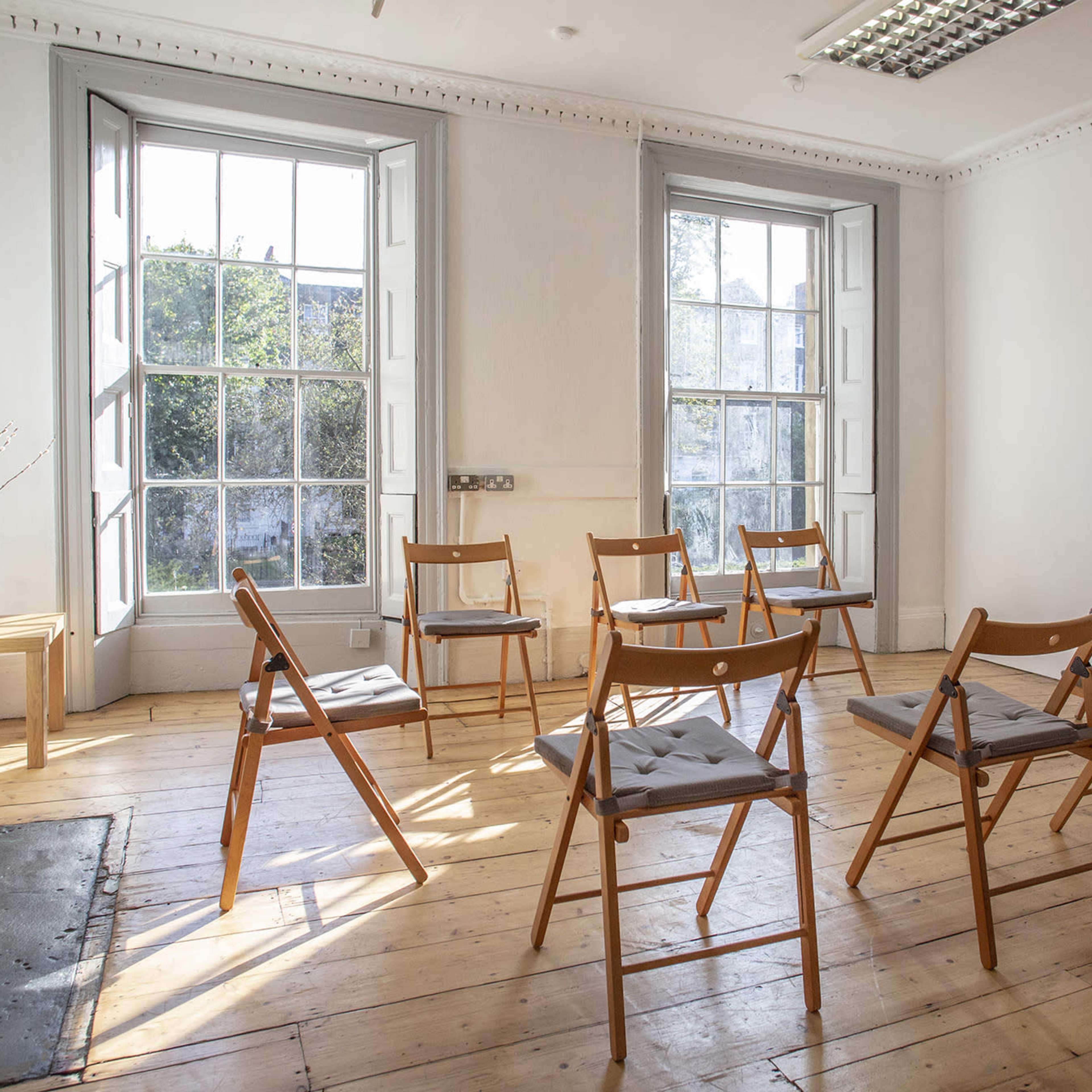 A round arrangement of wooden chairs is set up in a bright, minimally furnished room with large windows.