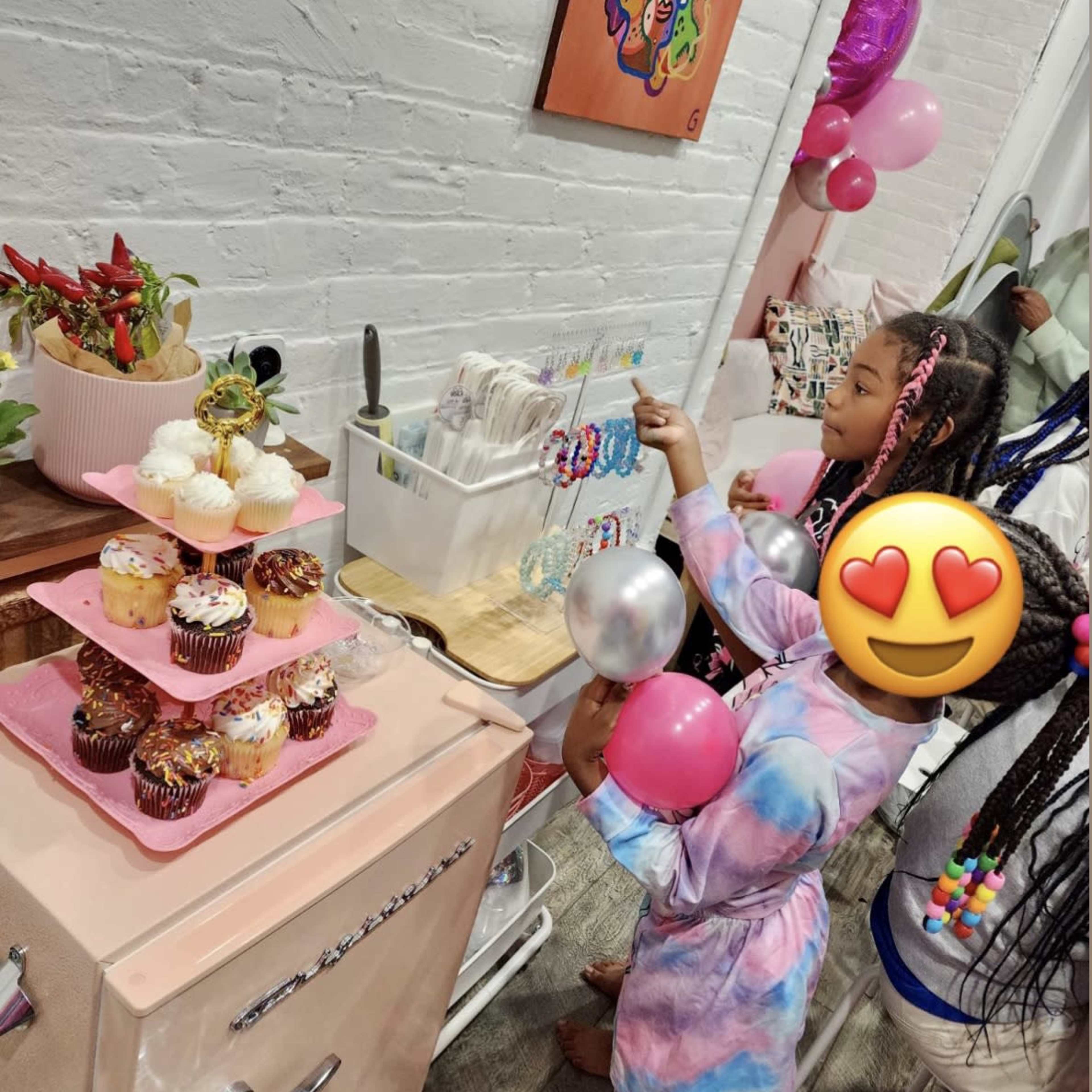 Two children are at a table decorated with cupcakes and balloons, one of them reaching for a treat while the other holds a pink balloon.
