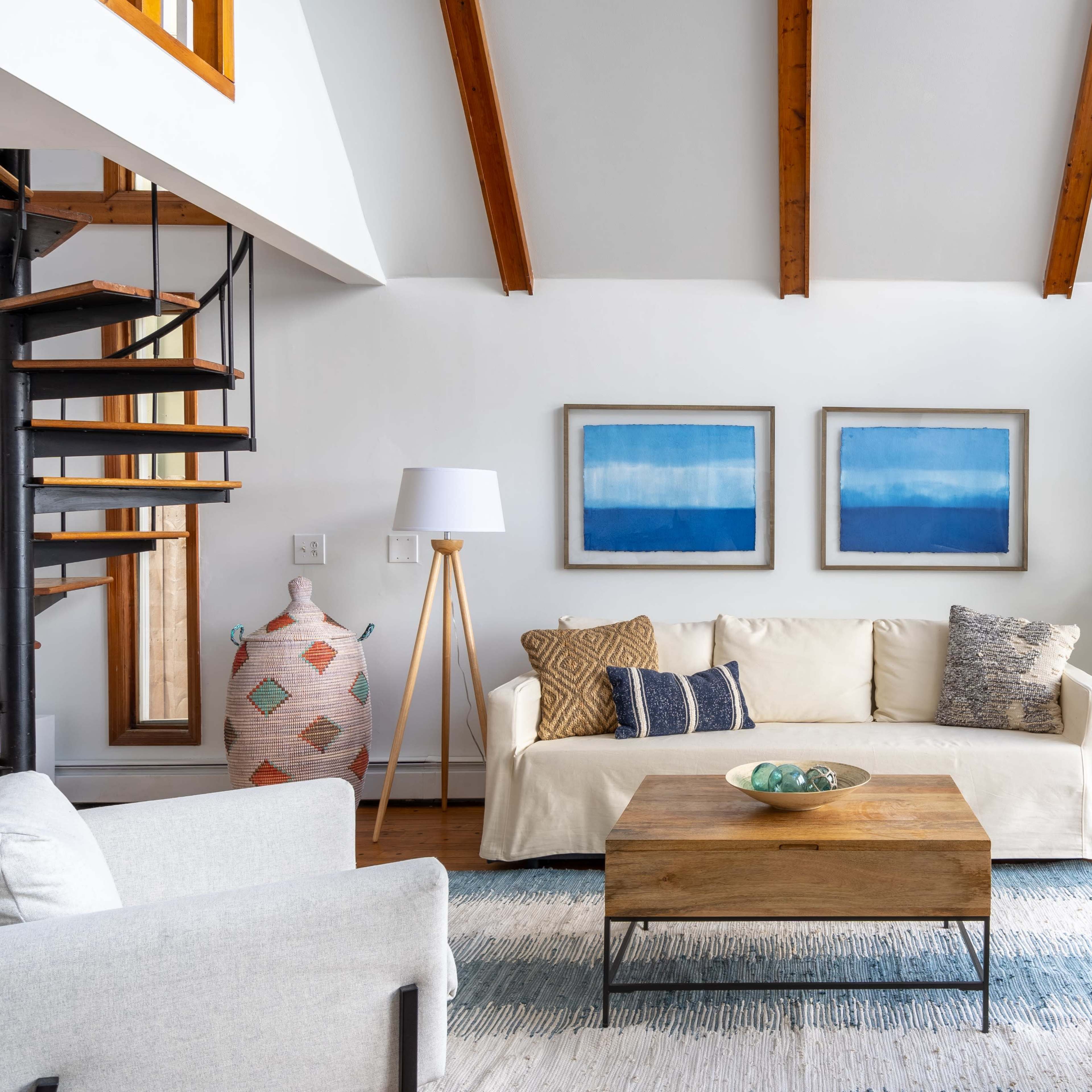 A living room features a light-colored sofa with decorative cushions, a wooden coffee table, two armchairs, and abstract artwork on the walls, alongside a spiral staircase and a large window.