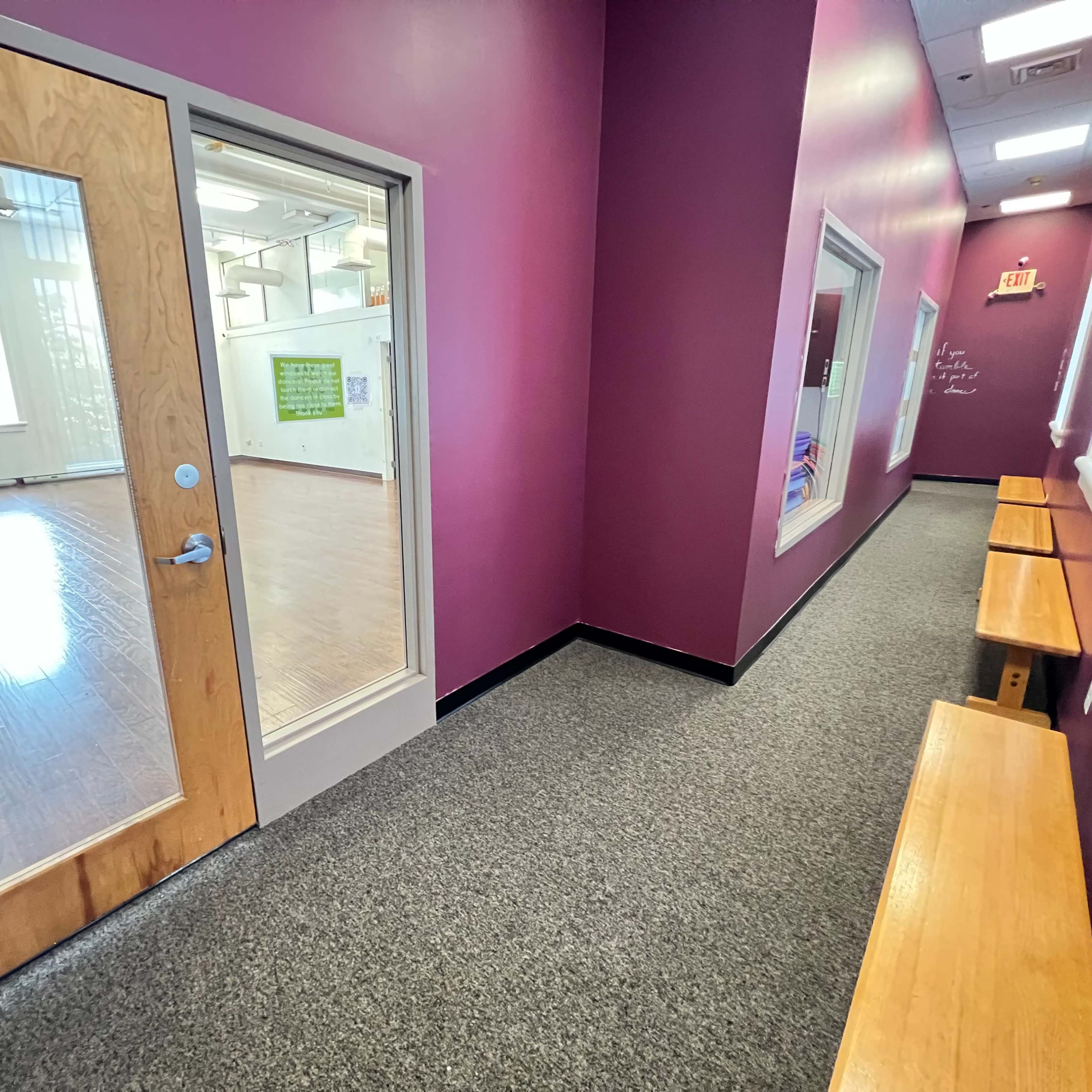 A narrow hallway with a purple wall, a glass door leading to a room with wooden flooring, and two benches along one side.