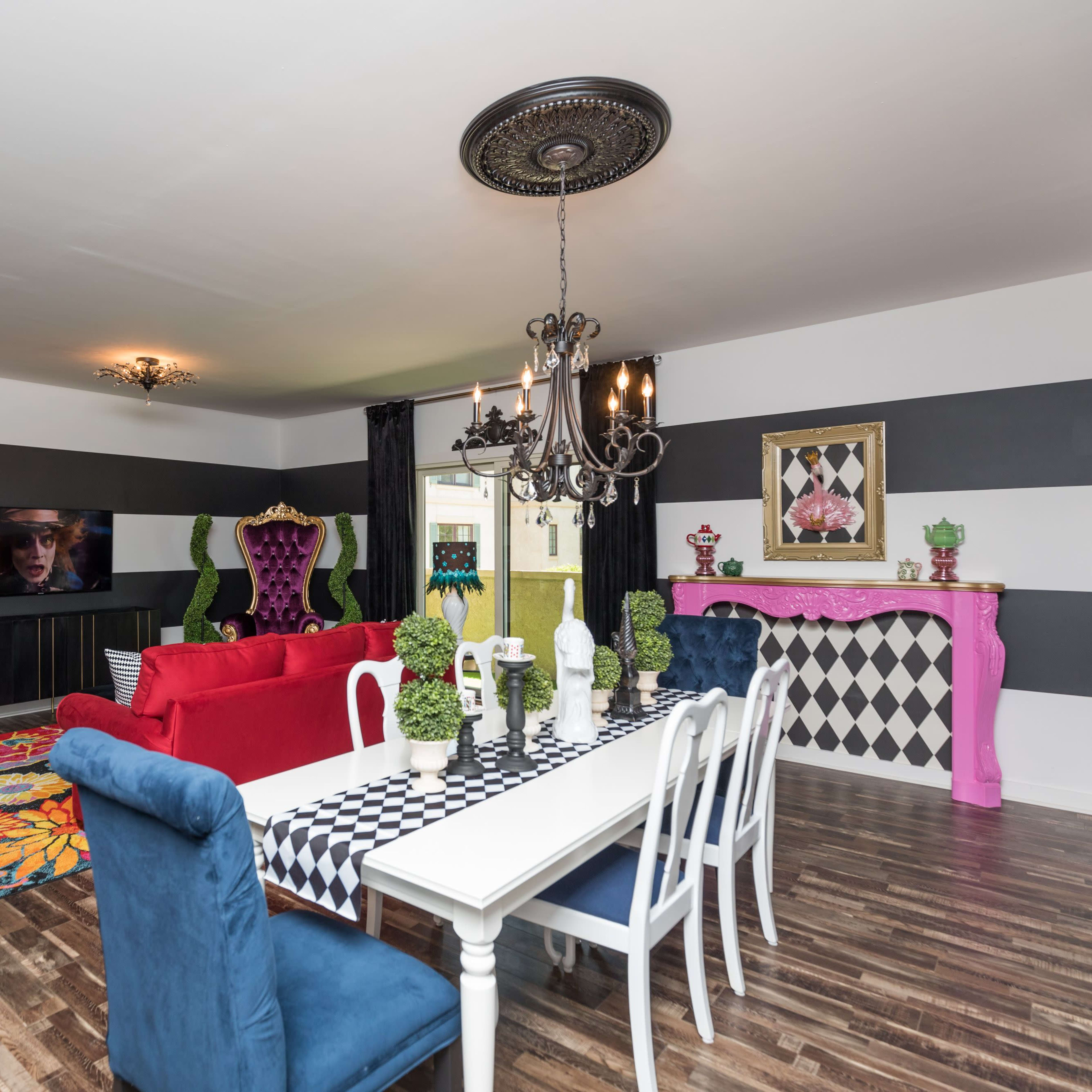 The living room features a mix of colorful furniture, including a red sofa and blue chairs, with striped walls, a large dining table, and various decorative elements.