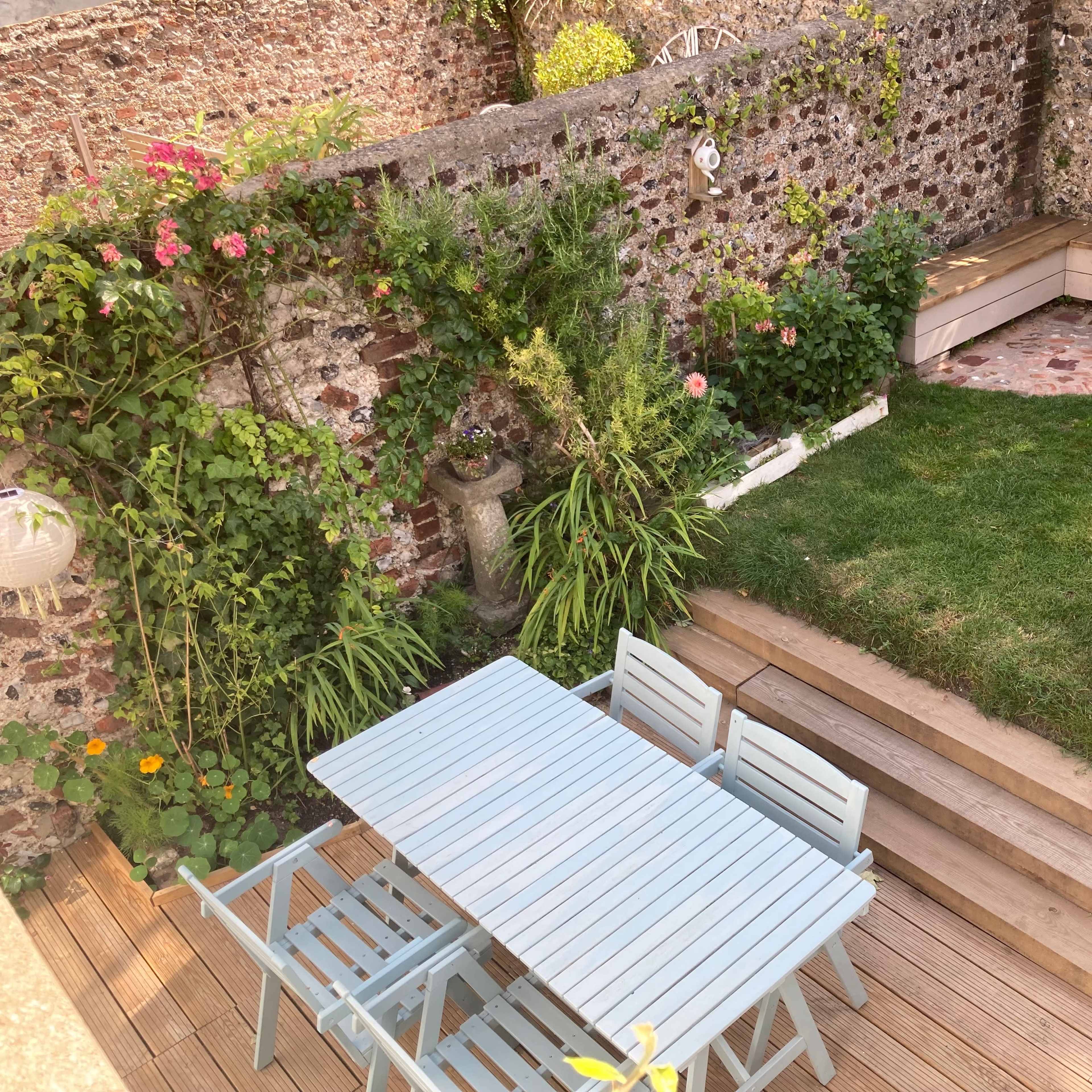 A well-maintained garden features a wooden dining table set with chairs, surrounded by green grass and flowering plants against a stone wall.