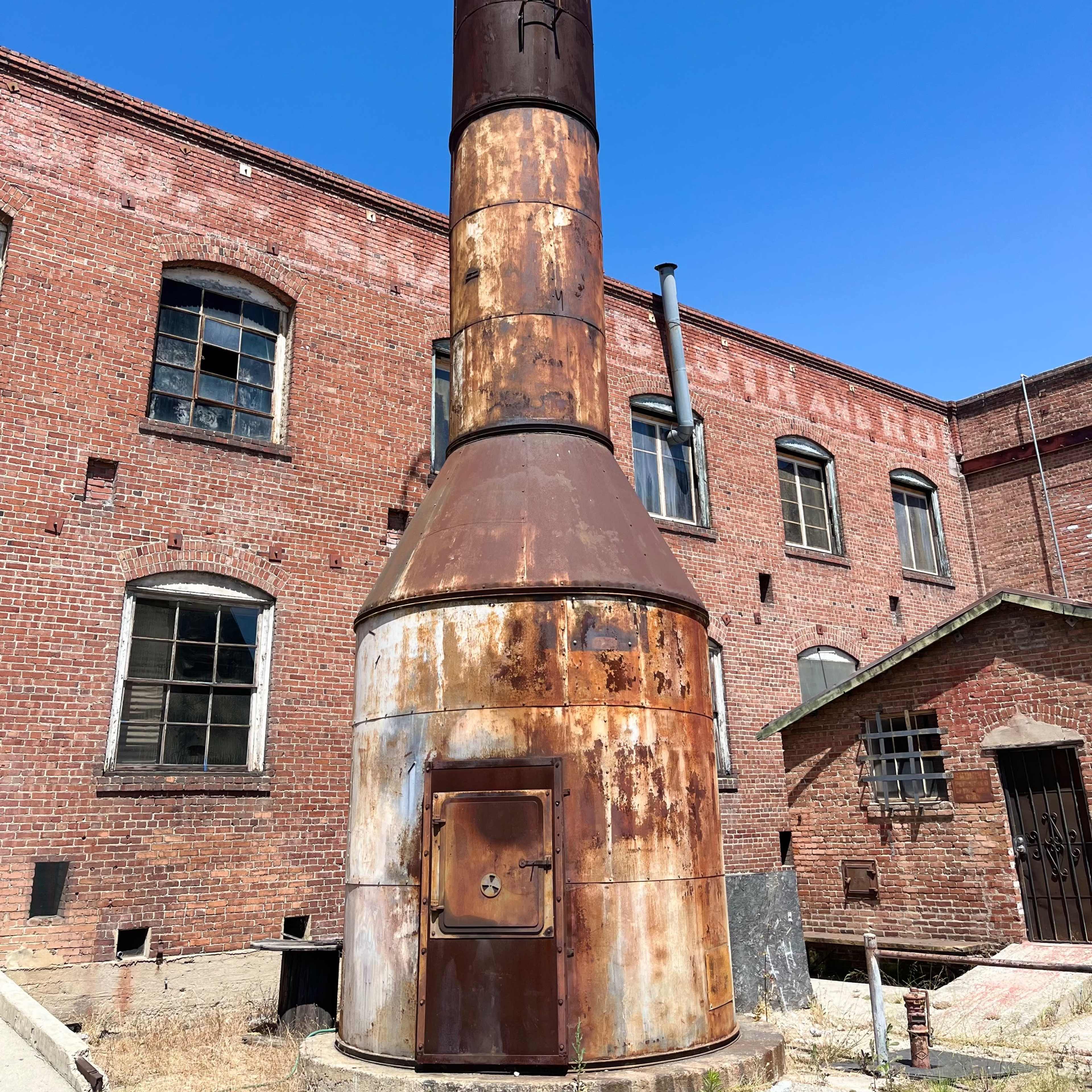 A tall, rusted industrial chimney stands next to a brick building under a clear blue sky.