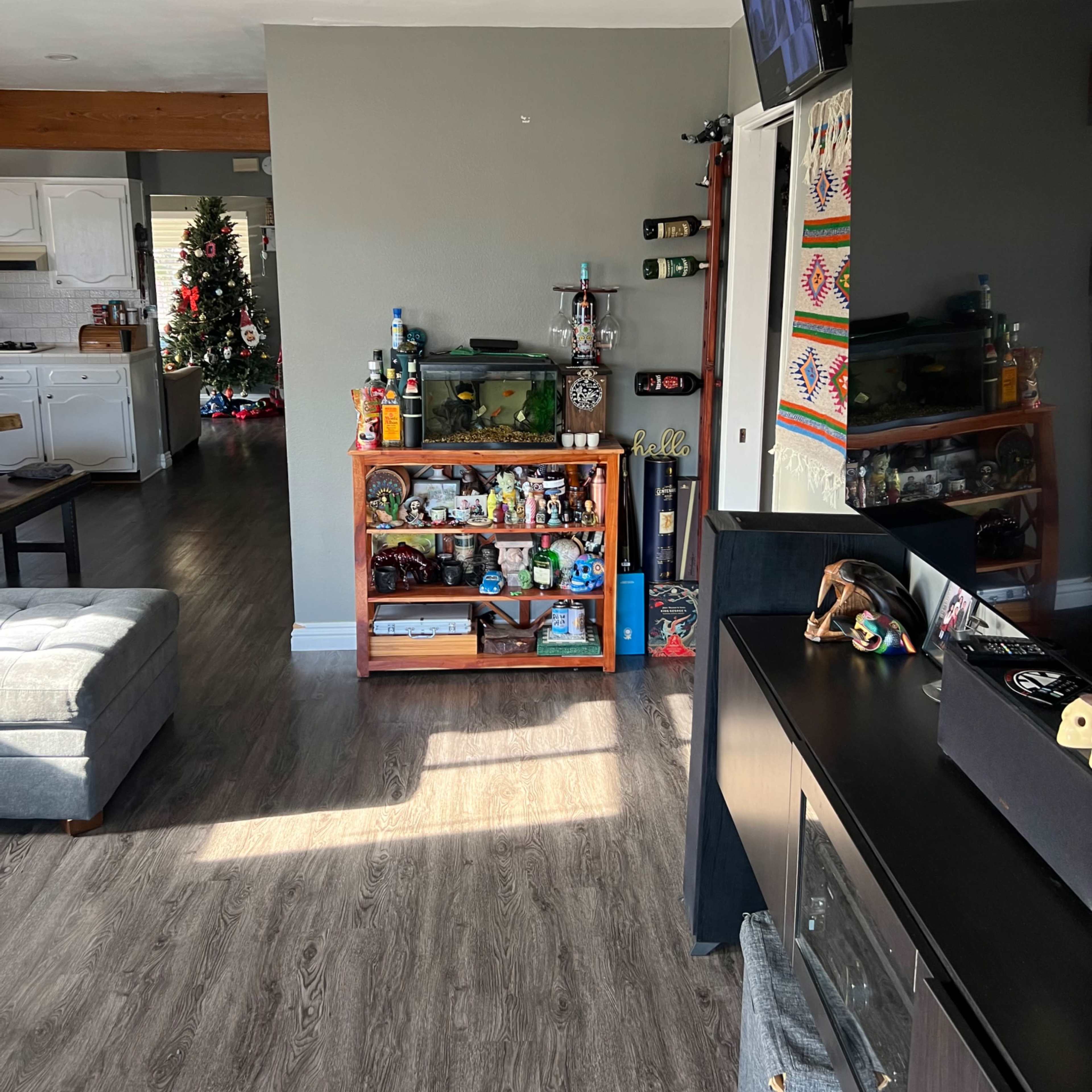 A living room area with a television mounted on the wall, a shelf displaying various items, and a glimpse of a kitchen with a Christmas tree in the background.