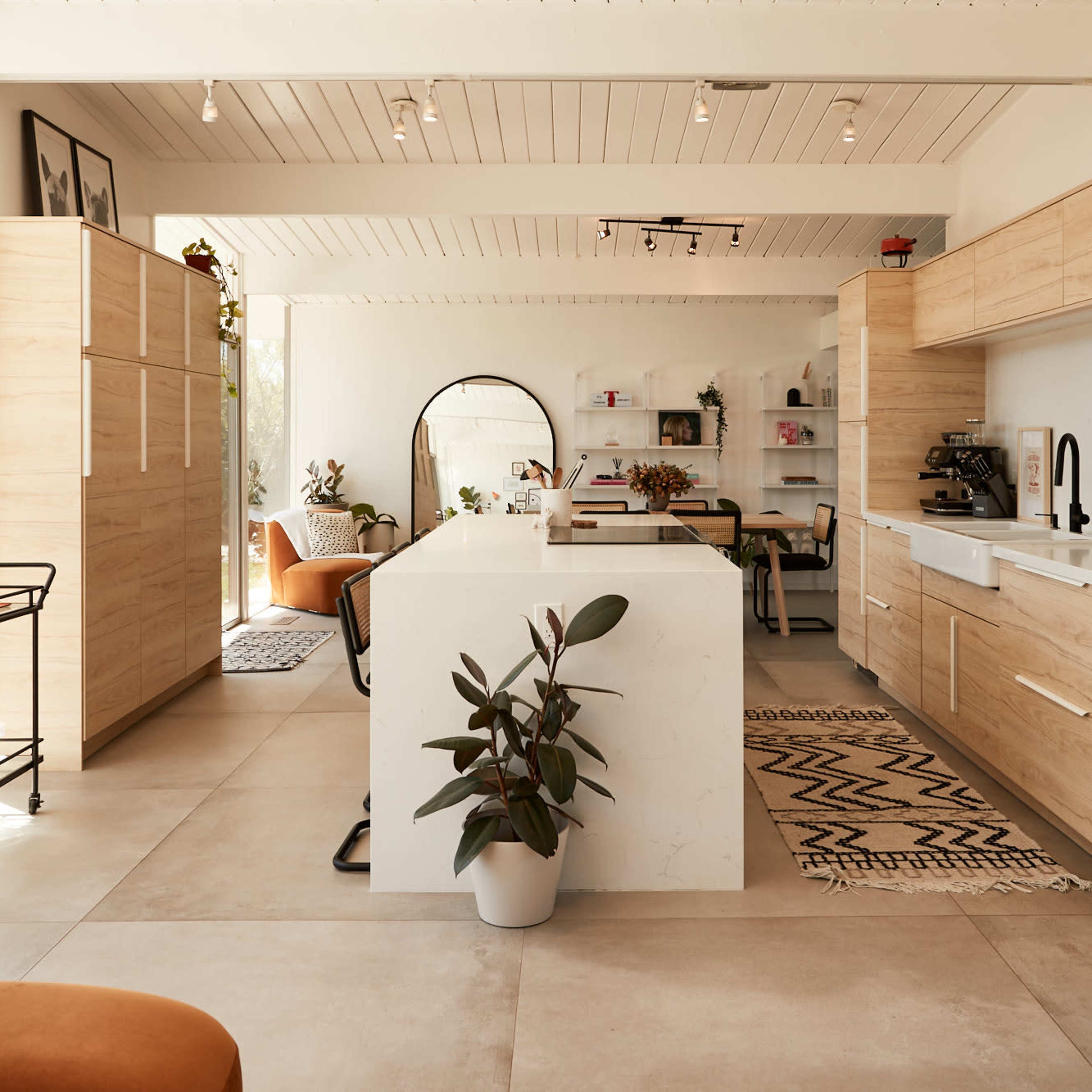 A modern kitchen featuring light wooden cabinetry, a central island, and a variety of indoor plants in a spacious, well-lit room.
