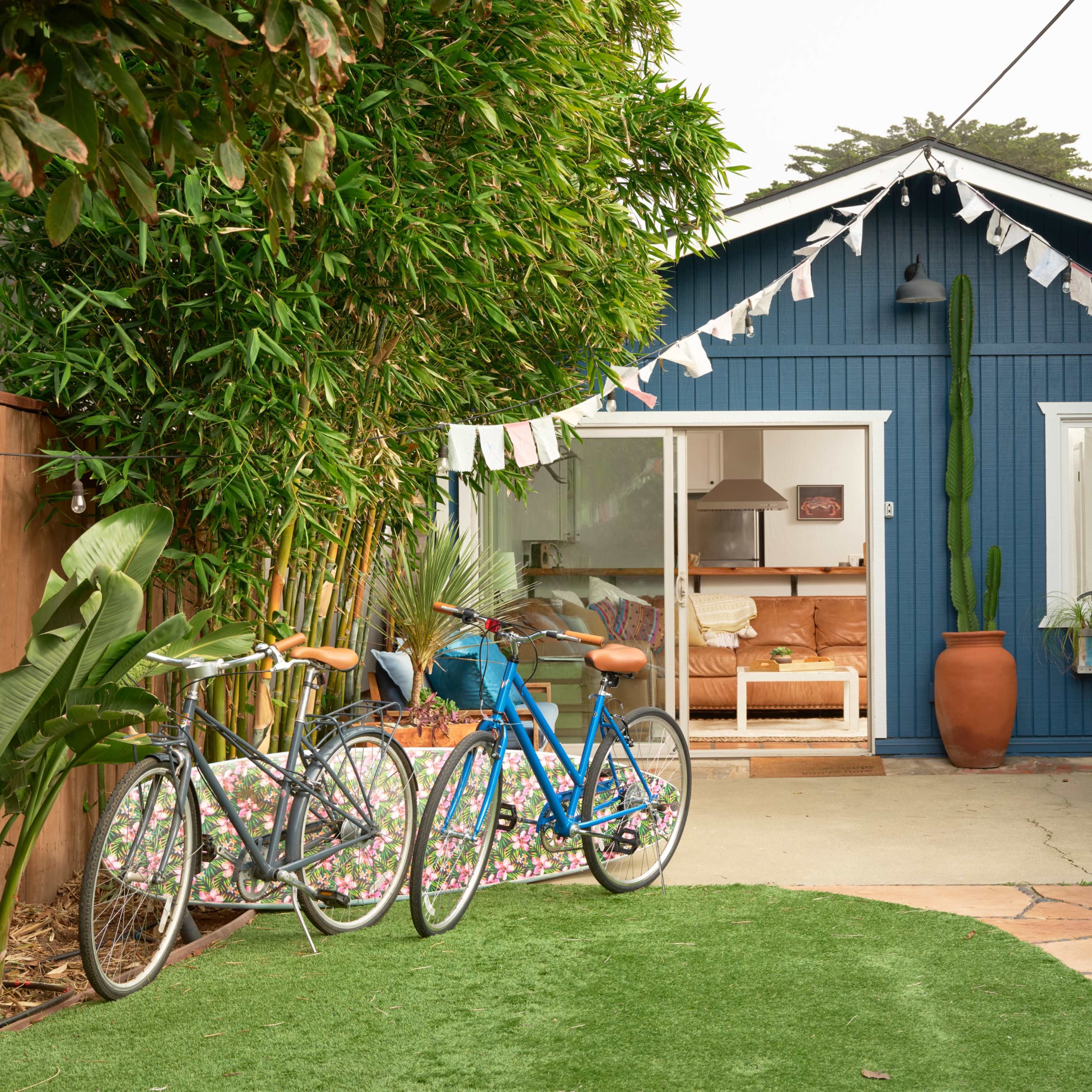 A blue cottage with an open front and a patio is surrounded by greenery and two bicycles are parked nearby.