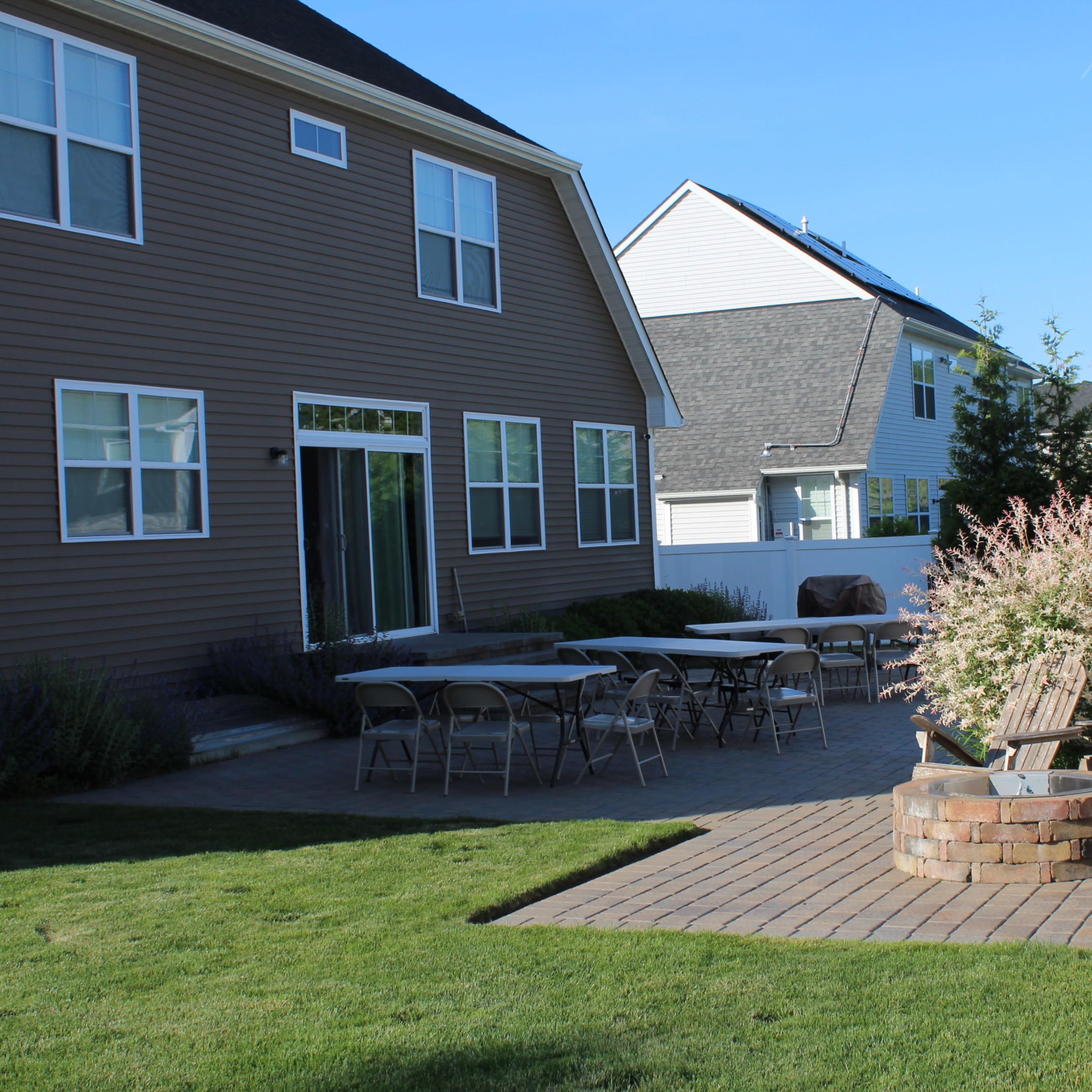 A backyard patio area with several tables and wooden chairs arranged around a circular stone fire pit, framed by green grass and shrubs.