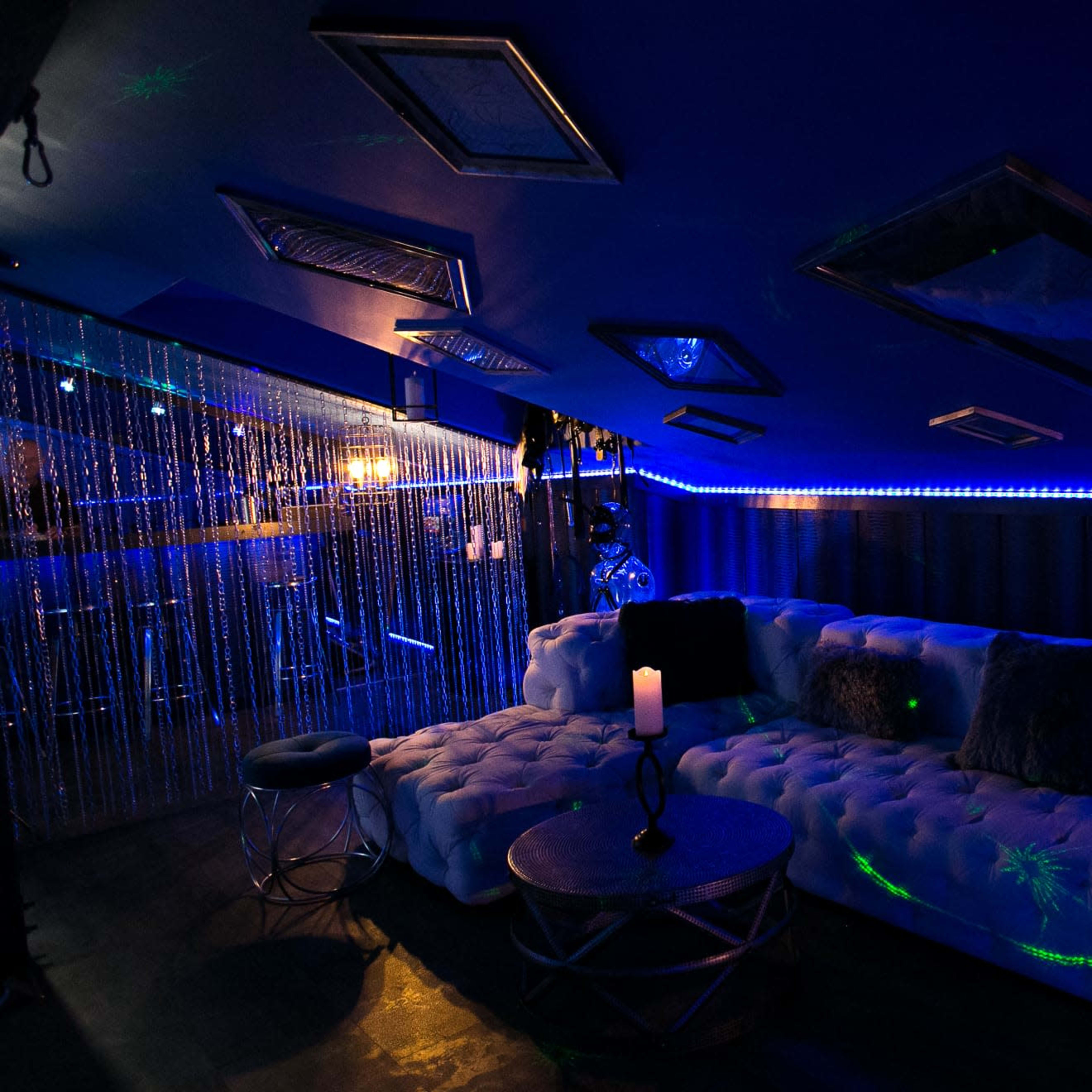 A dimly lit lounge area featuring a white tufted sectional sofa, a circular coffee table, and blue LED lighting along the walls.