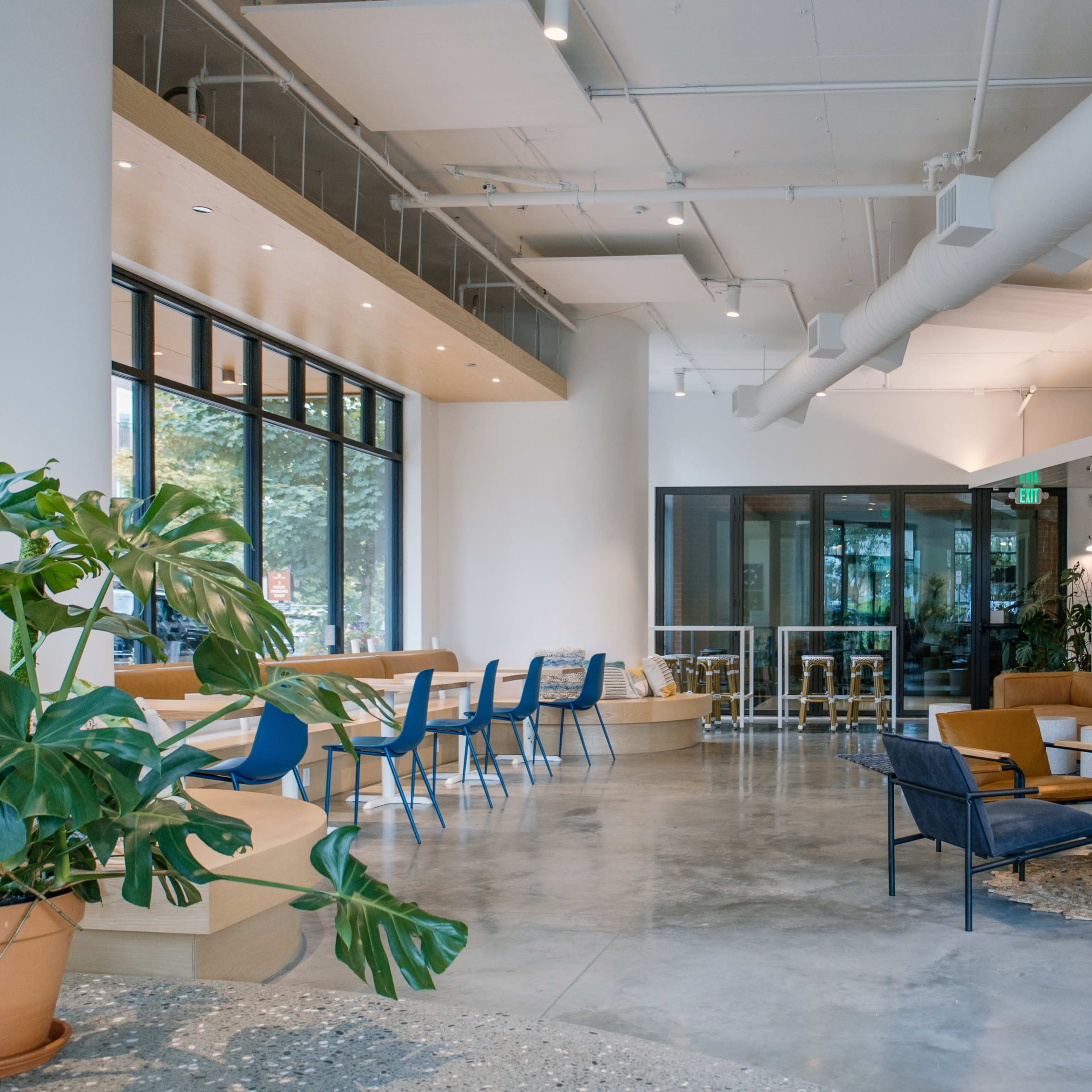 A modern, spacious interior of a café or lounge area with large windows, seating arrangements, and decorative plants.