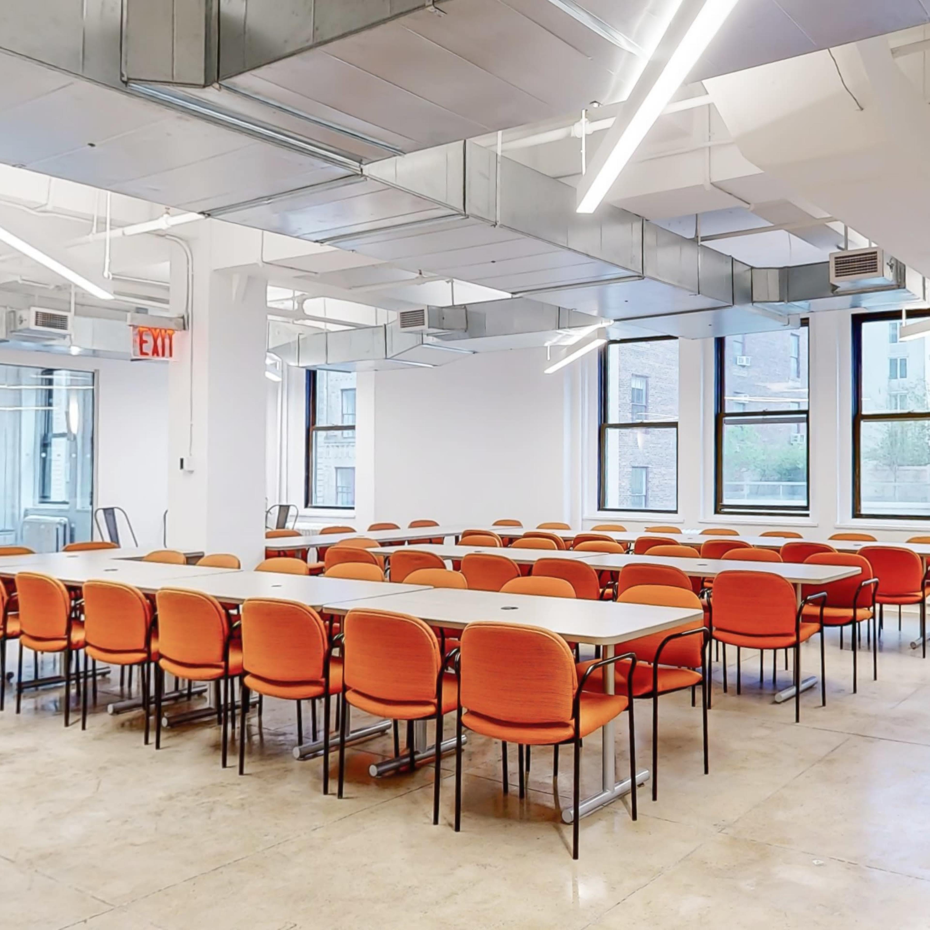 A spacious meeting room features rows of orange chairs arranged around white tables, with large windows allowing natural light to illuminate the space.