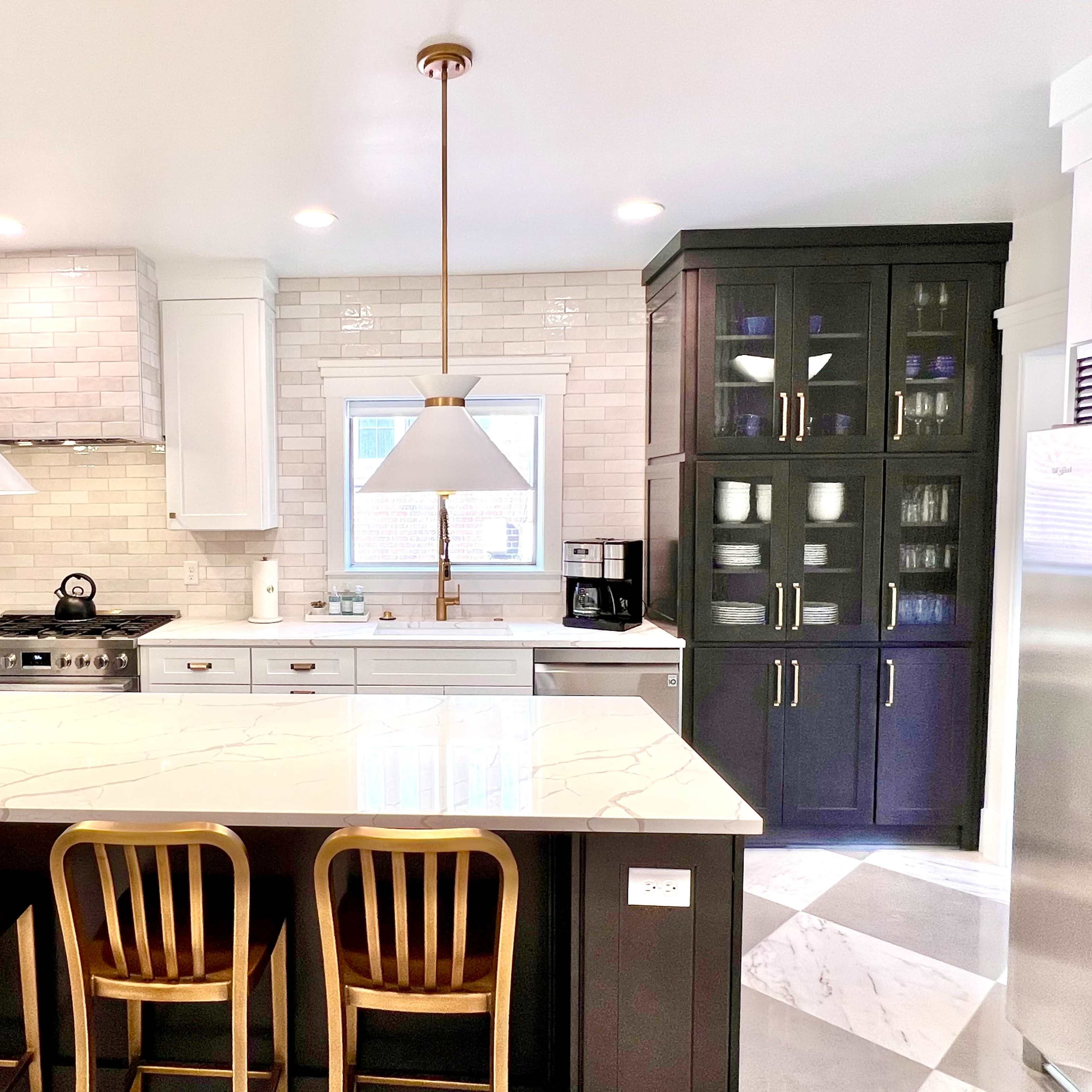 A modern kitchen features a central island with three bar stools, pendant lights above, and a combination of dark cabinetry and white tile backsplash.