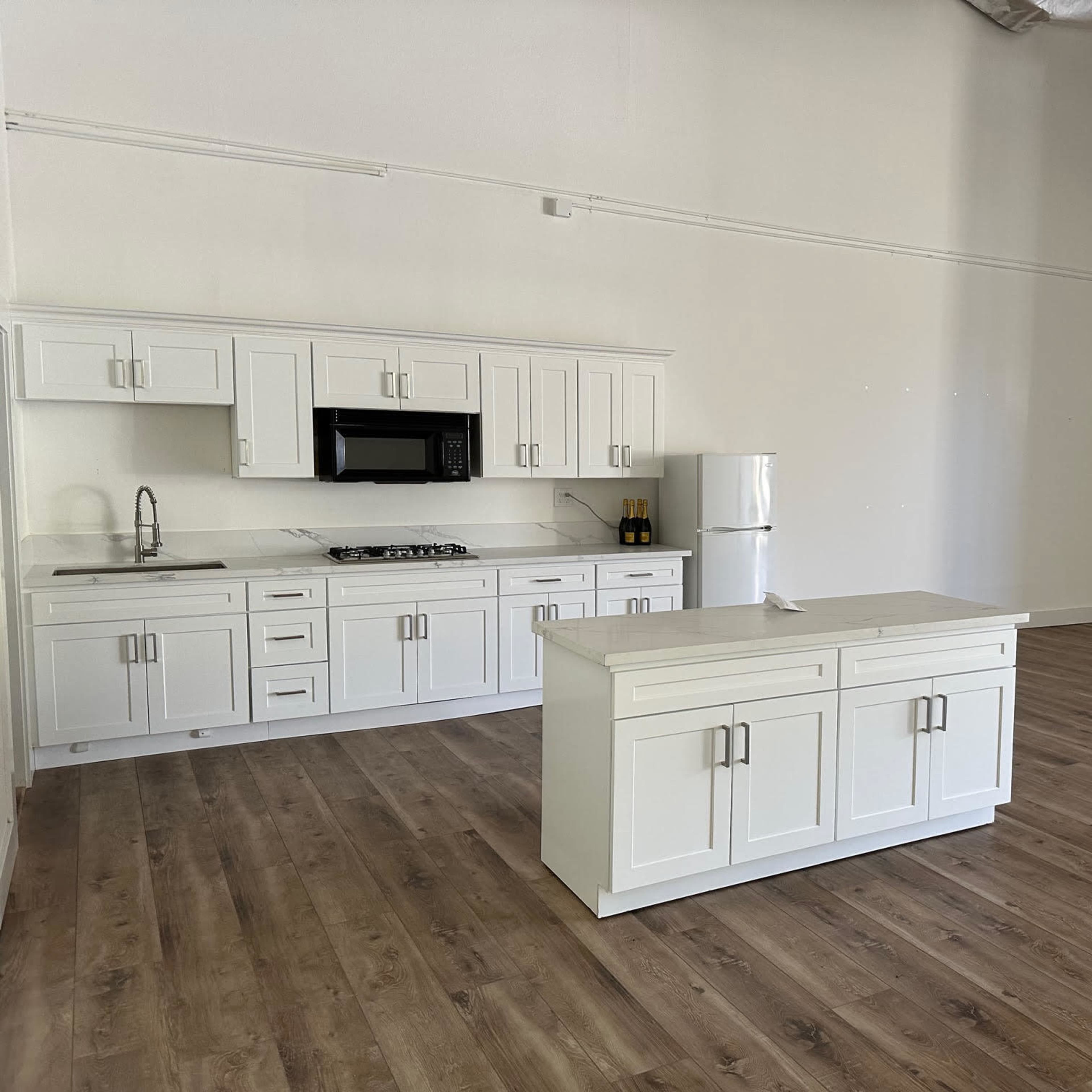 A modern kitchen with white cabinets, a refrigerator, a microwave, and a central island on a wooden floor in a spacious, empty room.