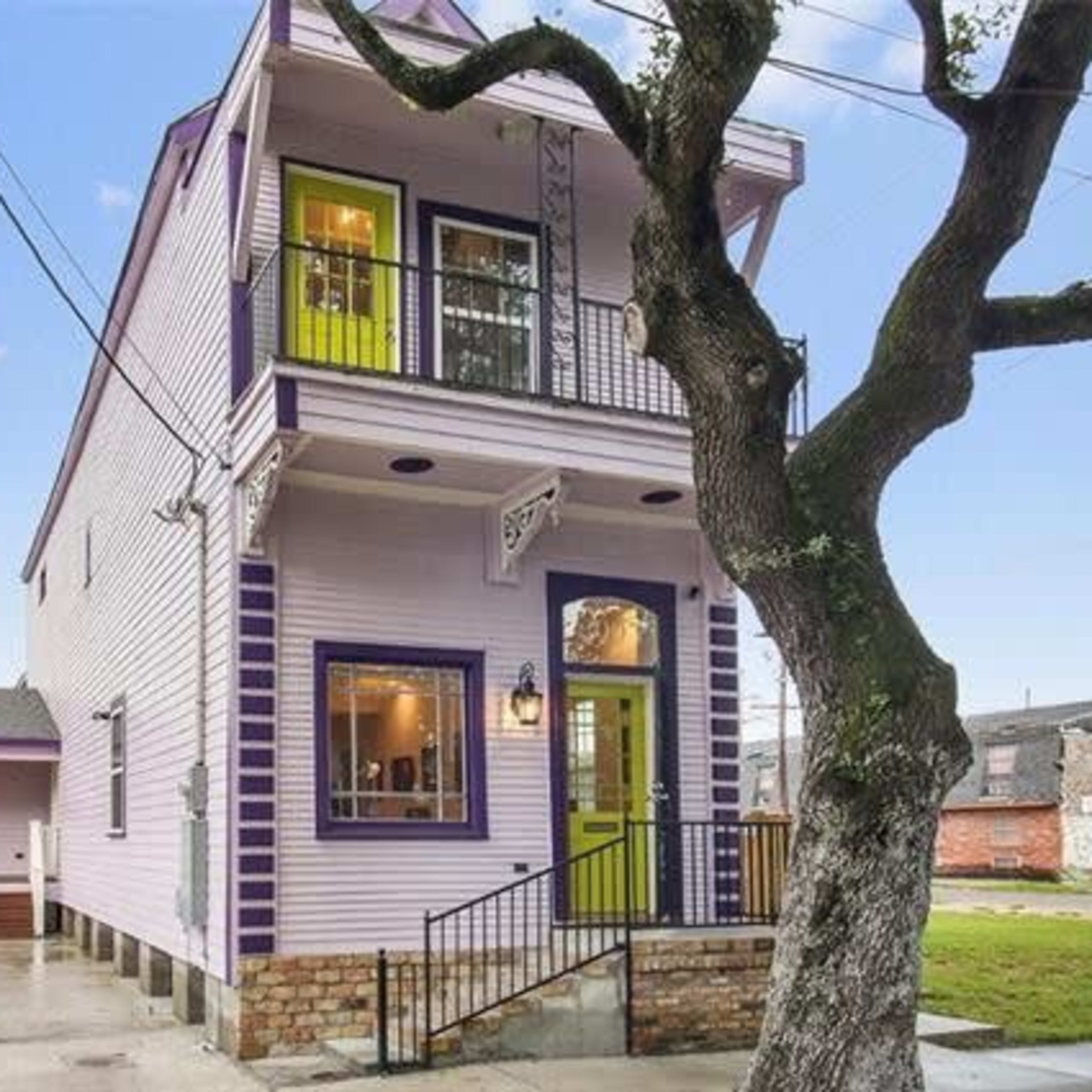 A three-story, purple and white house with green accents, featuring a porch and a tree in the front yard.