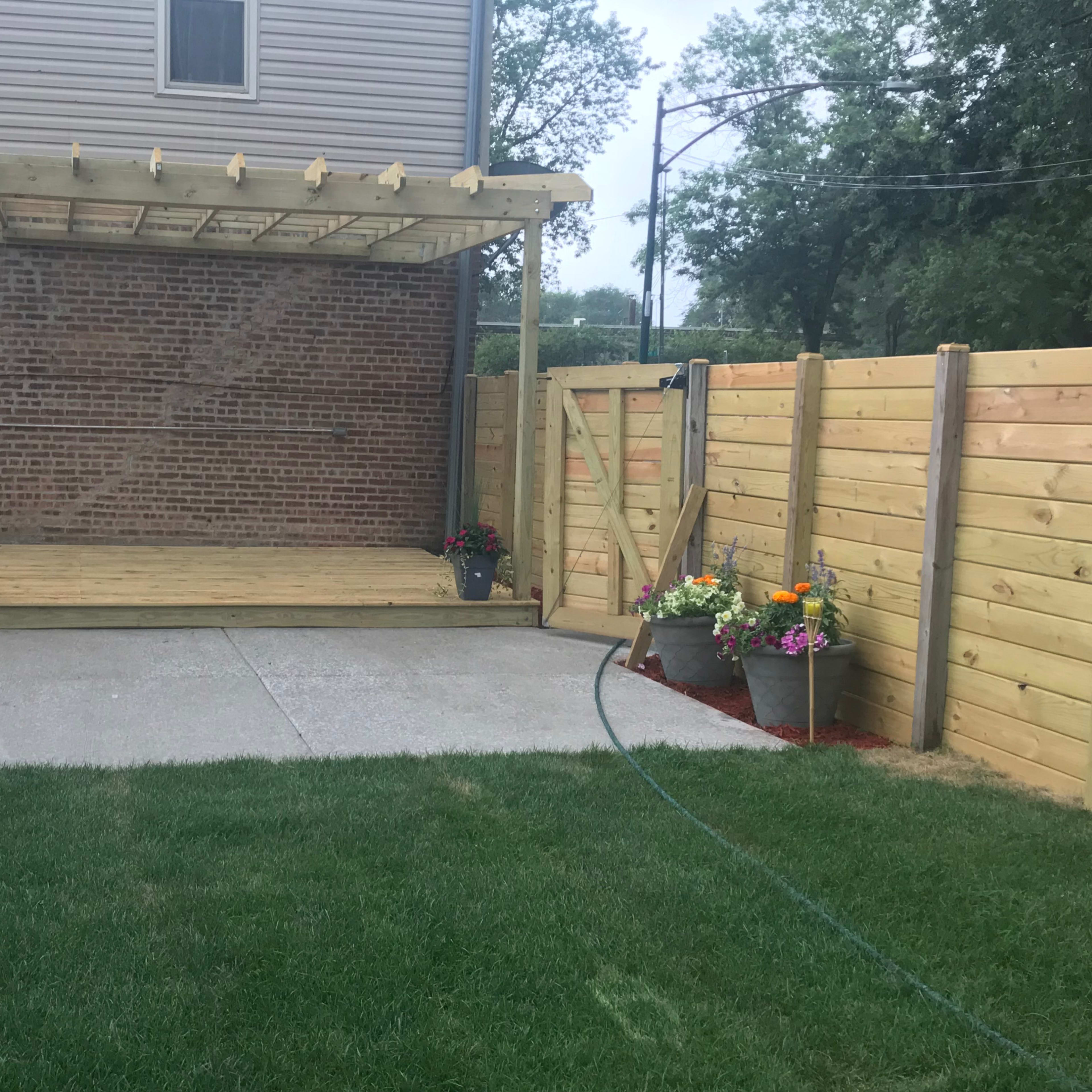 A wooden deck with a pergola, surrounded by a fenced yard with flower pots and a grassy area.