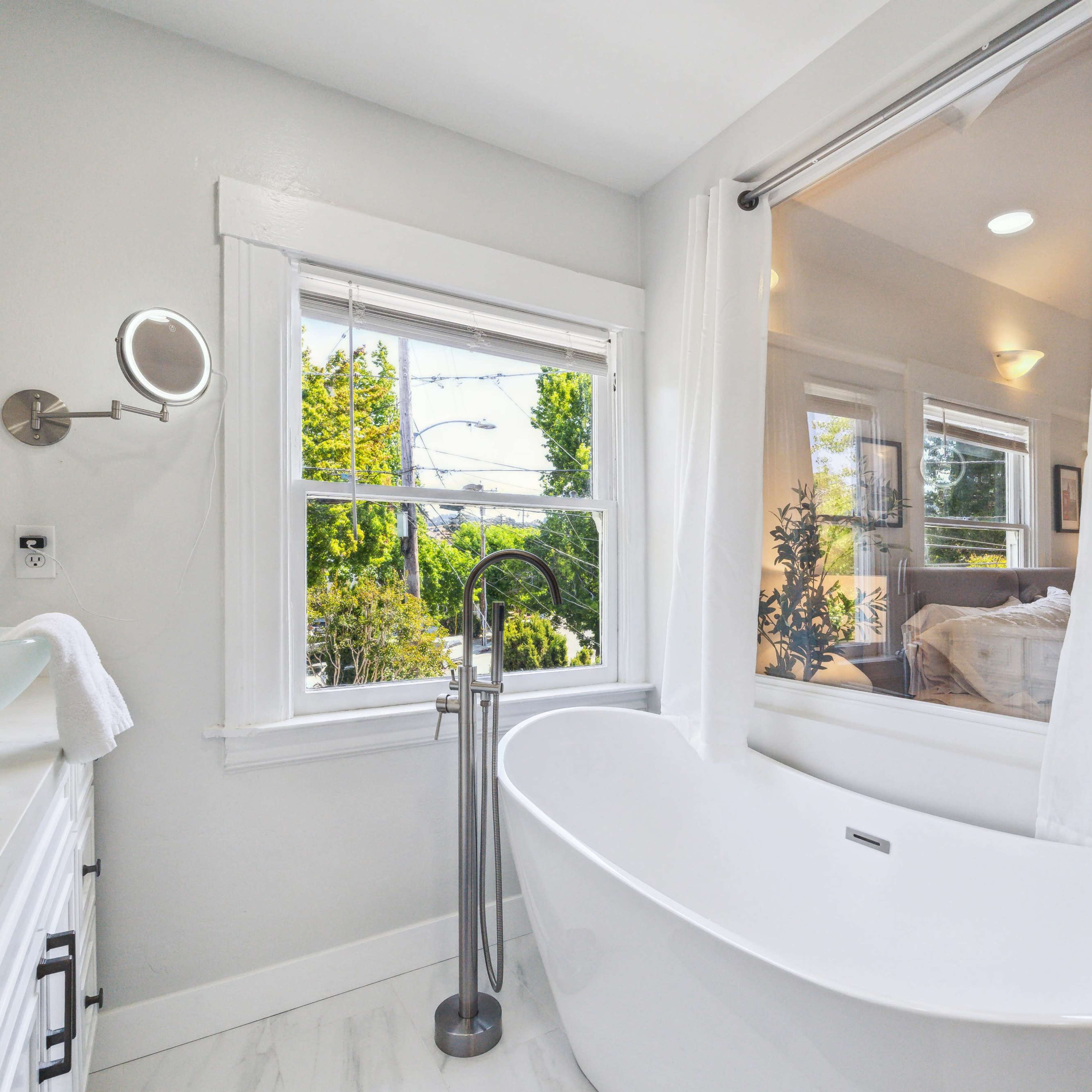 A modern bathroom featuring a freestanding white bathtub beside a window with a view of greenery, white cabinetry, and a wall-mounted mirror.
