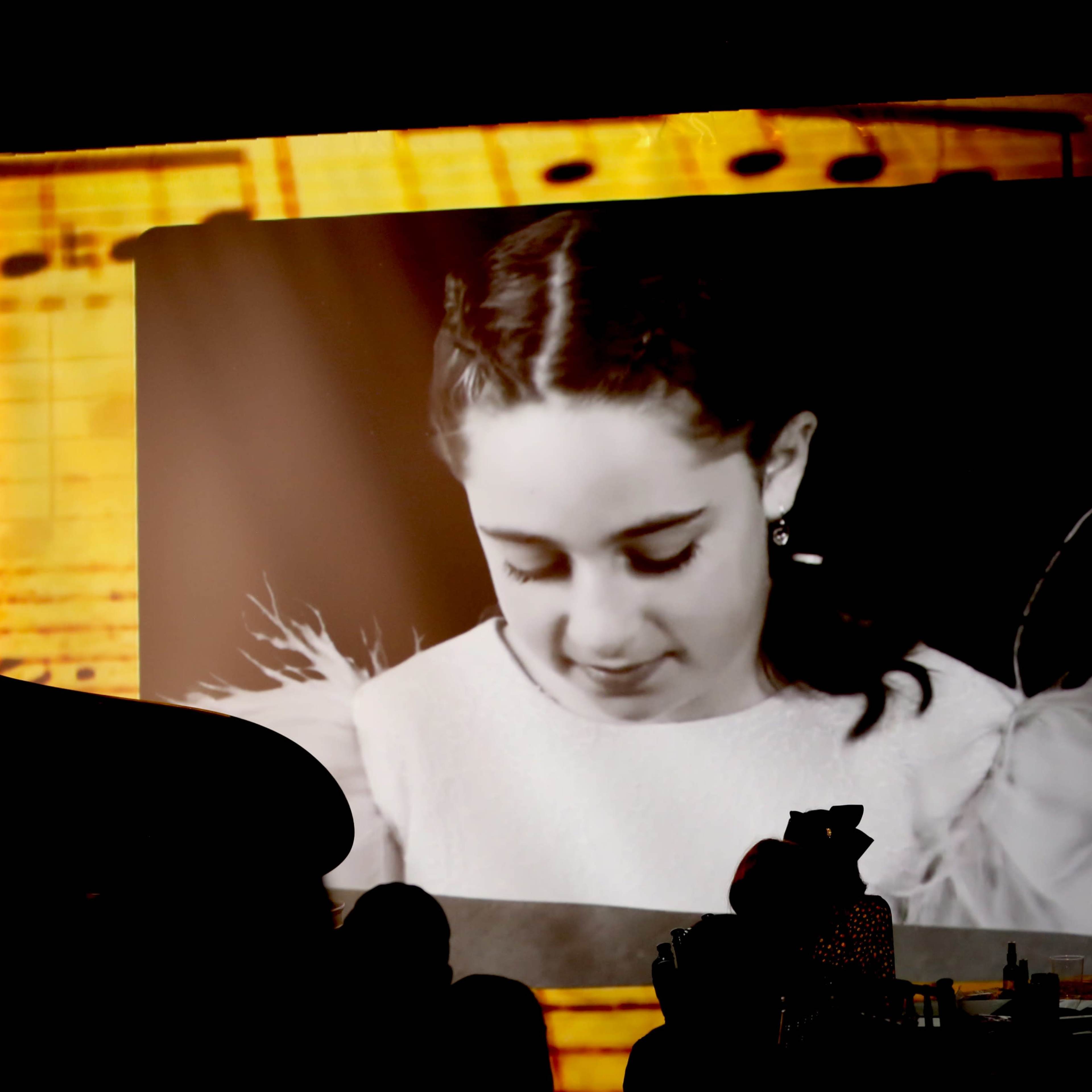 A silhouette of a person sits at a piano in front of a large projected image of a young girl looking downward, with a backdrop featuring musical notes.