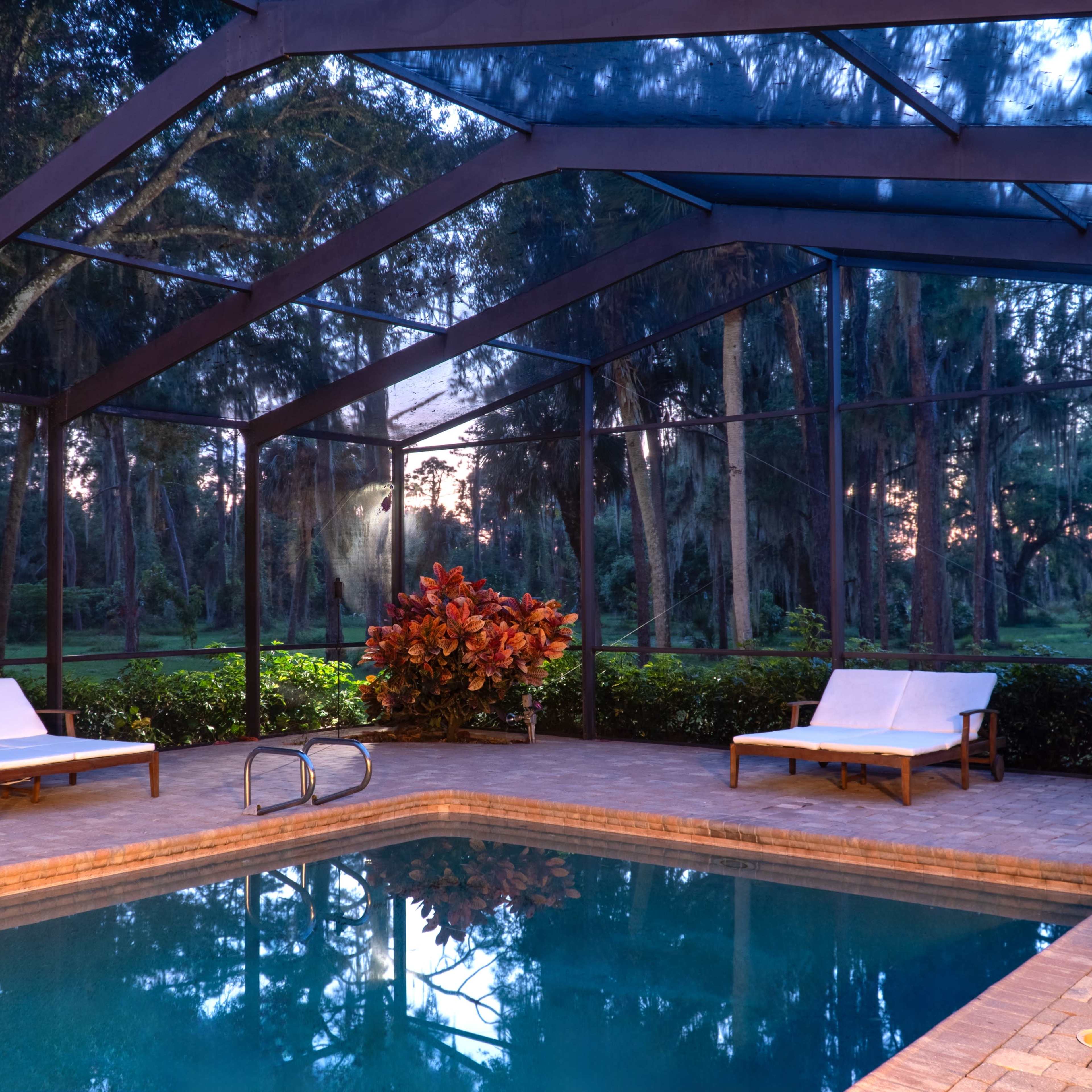 The scene shows a screened-in pool area at dusk with two lounge chairs beside the water and lush greenery surrounding the space.