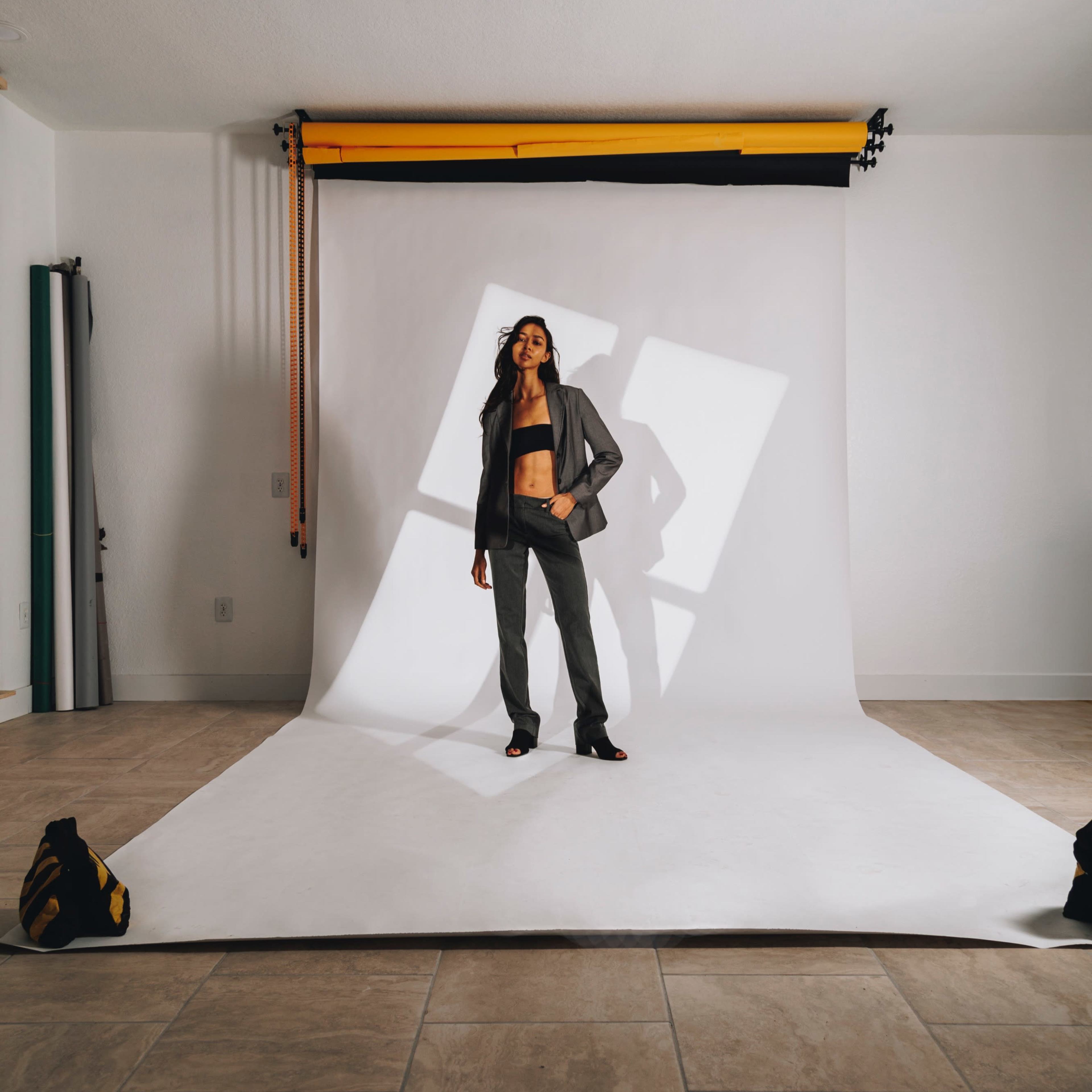 A model poses on a white backdrop in a studio, surrounded by studio equipment and lighting fixtures.