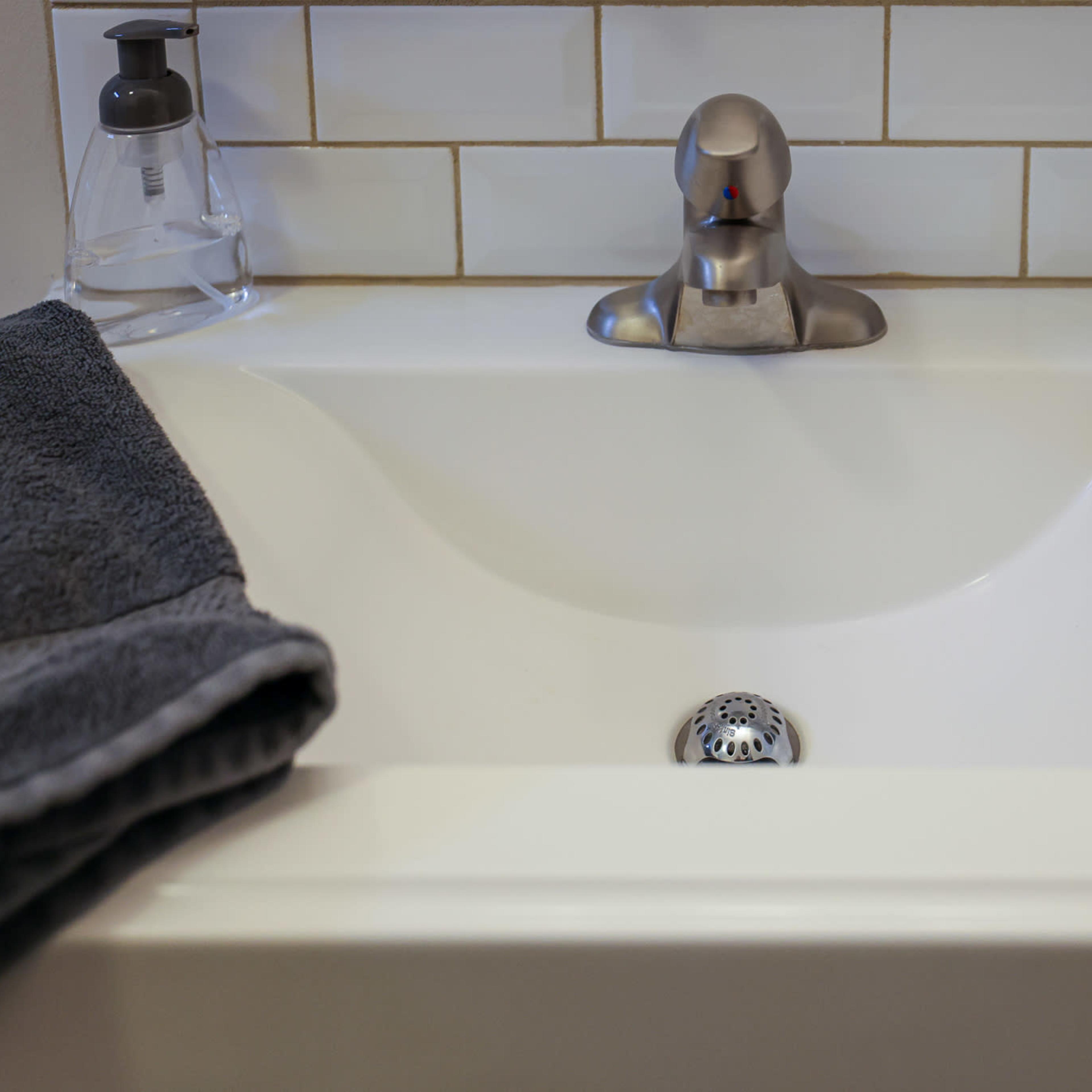 A white sink with a stainless steel faucet and a black cup sits against a tiled backsplash, accompanied by a gray towel.