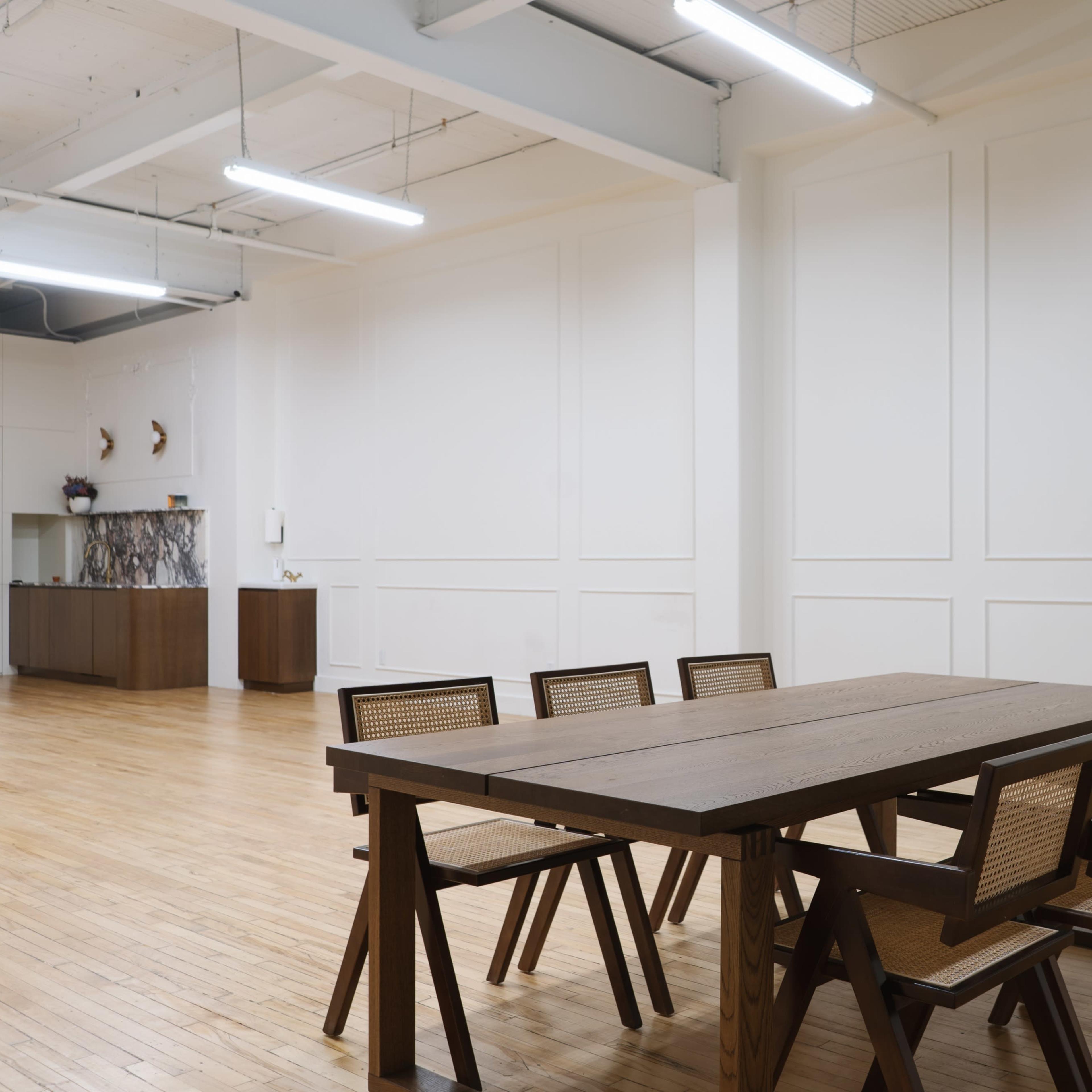 A spacious room features a large wooden table surrounded by six chairs, with plain white walls and a minimalist design.