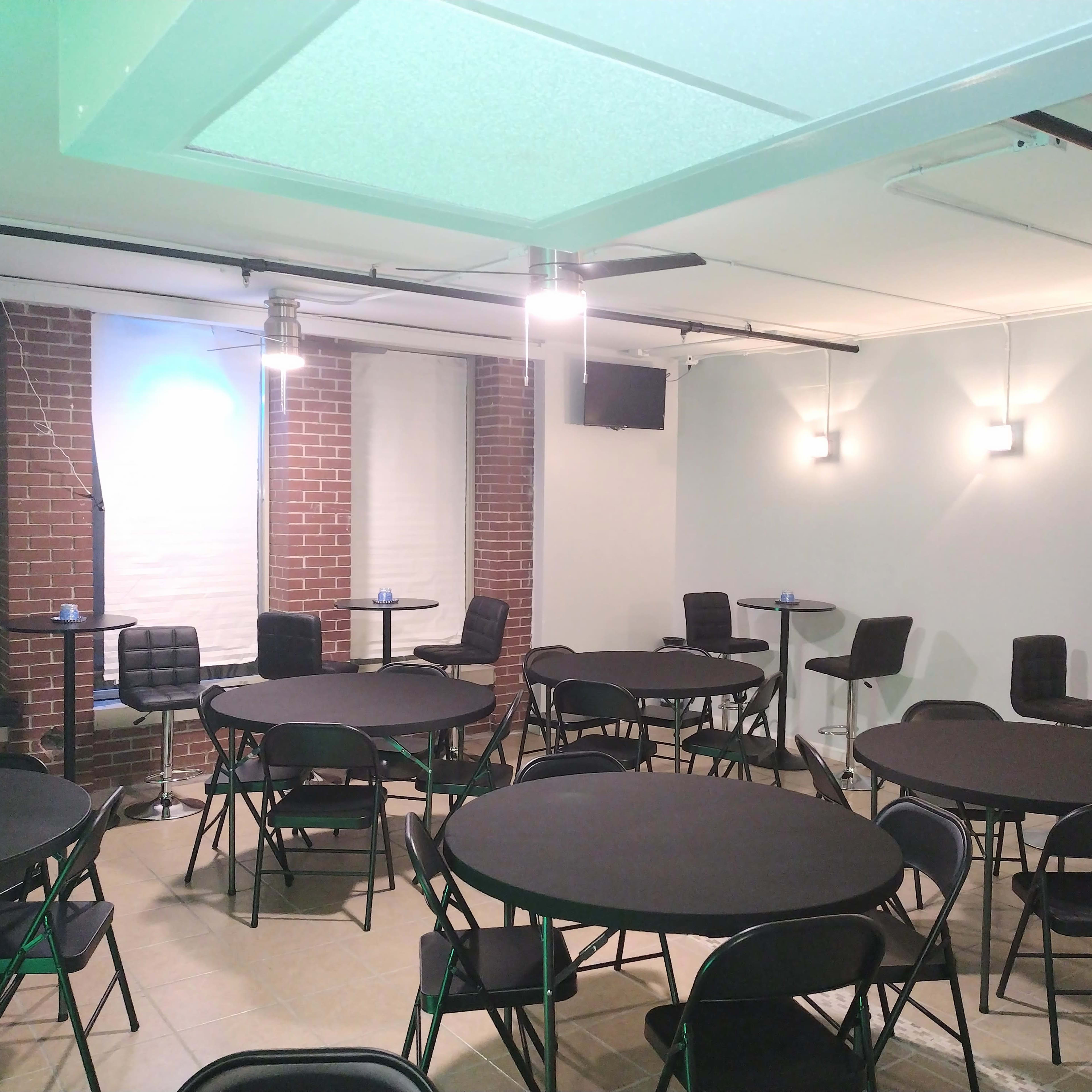A room set up for an event, featuring several round black tables surrounded by folding chairs, with brick walls and modern lighting.
