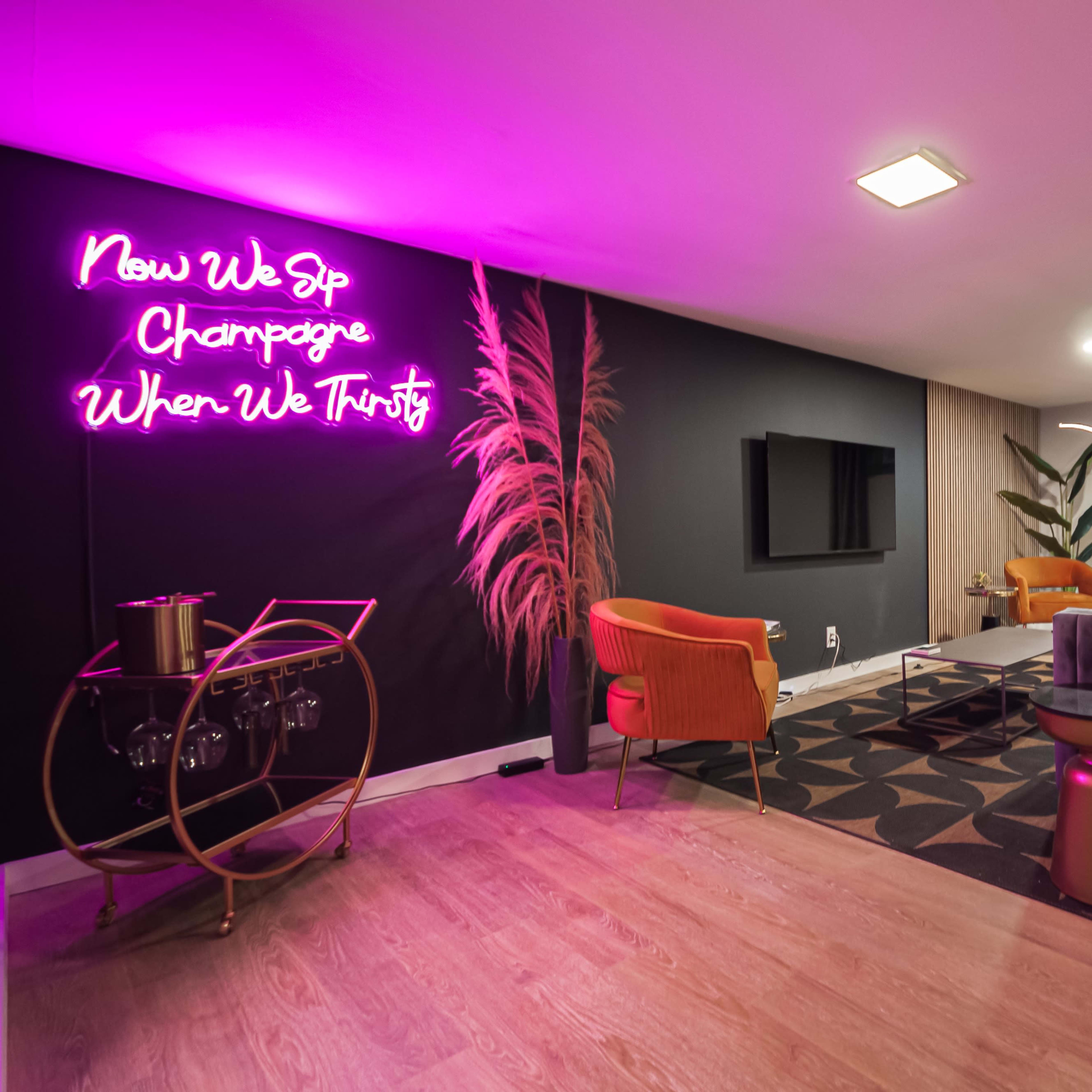 A modern living room features a gray velvet sofa, two orange chairs, a round table, and a neon sign on the wall that reads, "Now We Sip Champagne When We Thirsty."