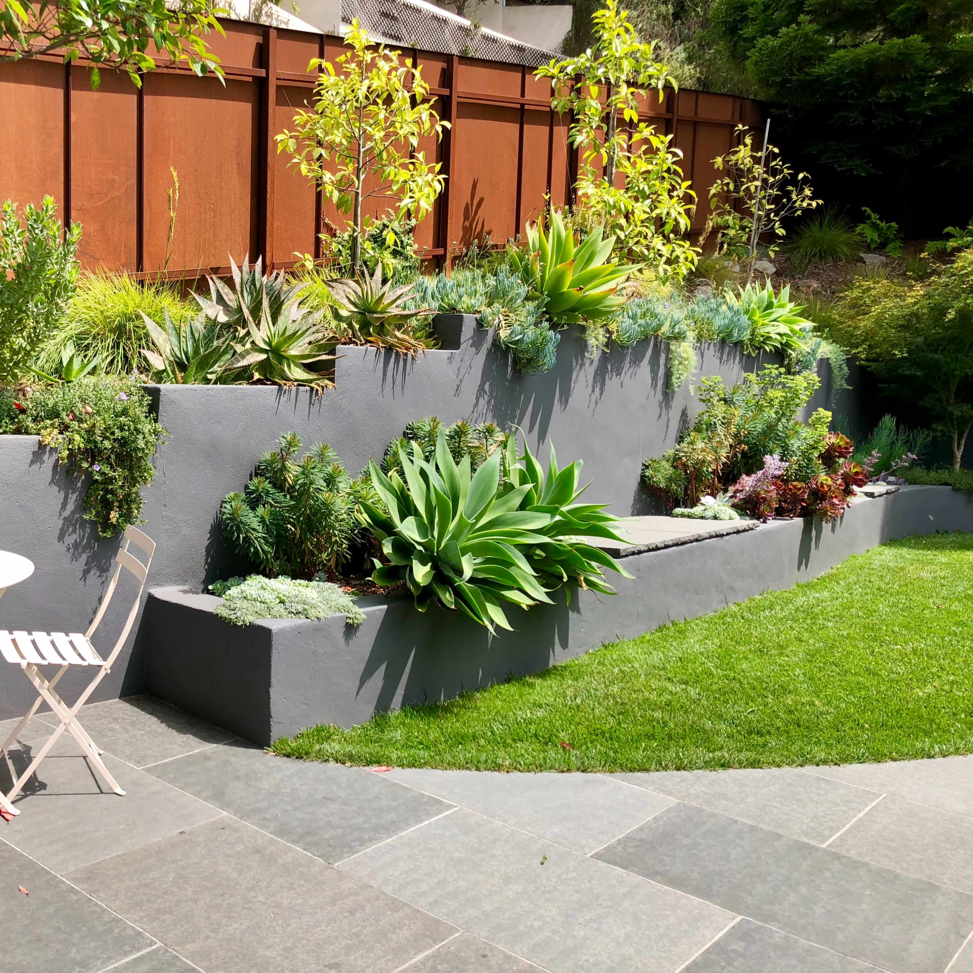 A landscaped garden with layered planter boxes filled with various plants, alongside a small round table and chairs on a stone patio.