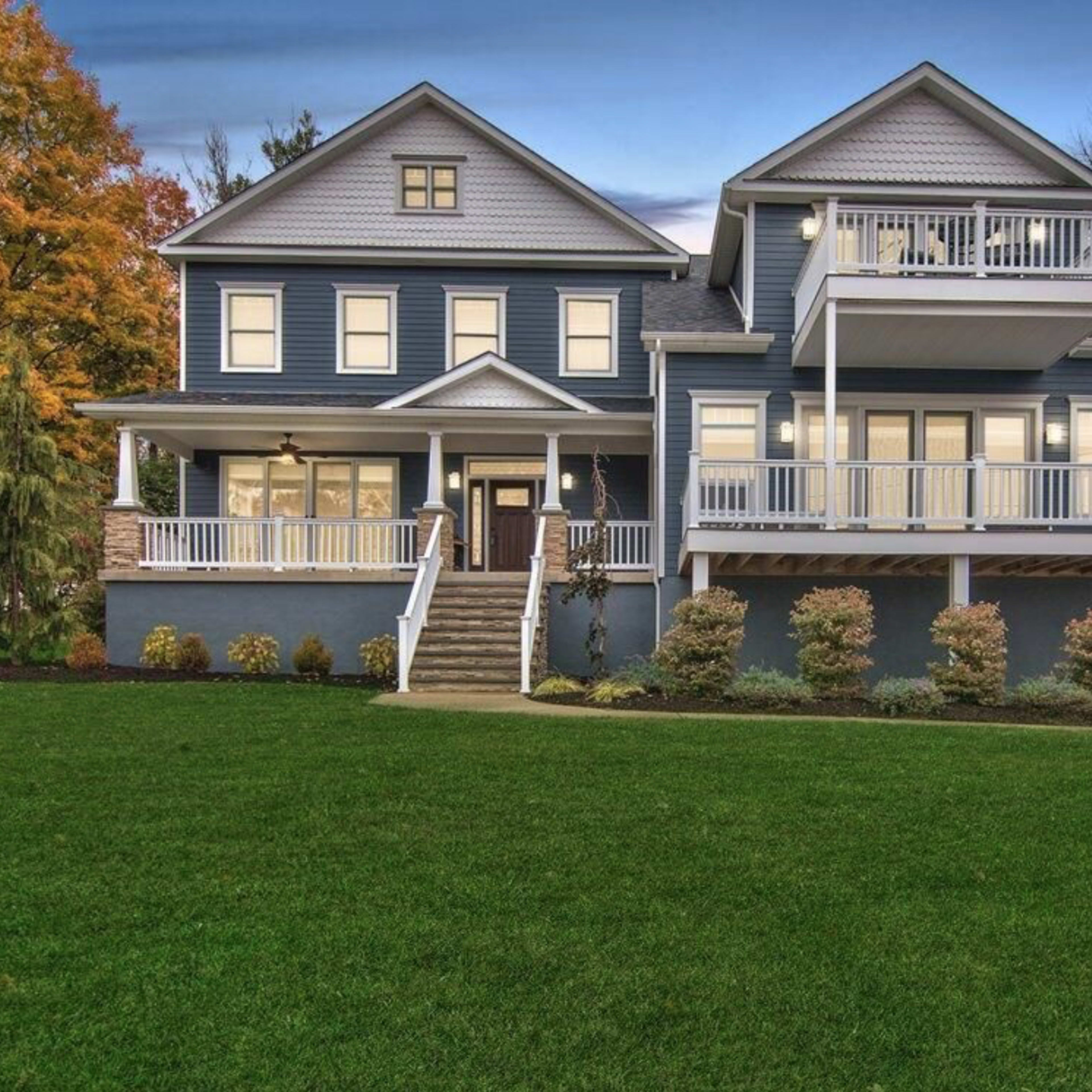 A large, two-story blue house with a white porch and multiple balconies, surrounded by a neatly manicured lawn and trees in autumn foliage.