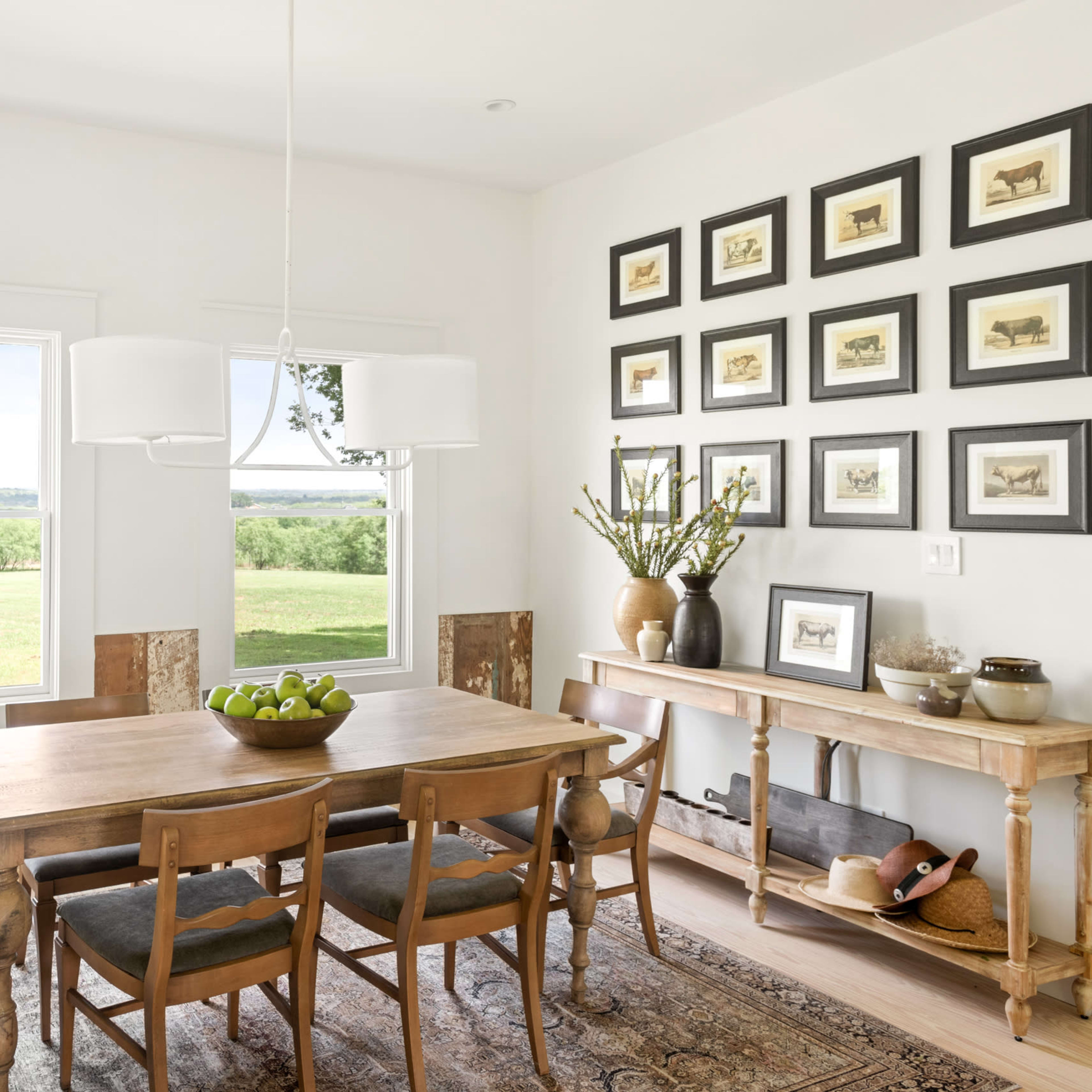 A bright dining room features a wooden table with four chairs, surrounded by large windows and framed artwork on the walls.