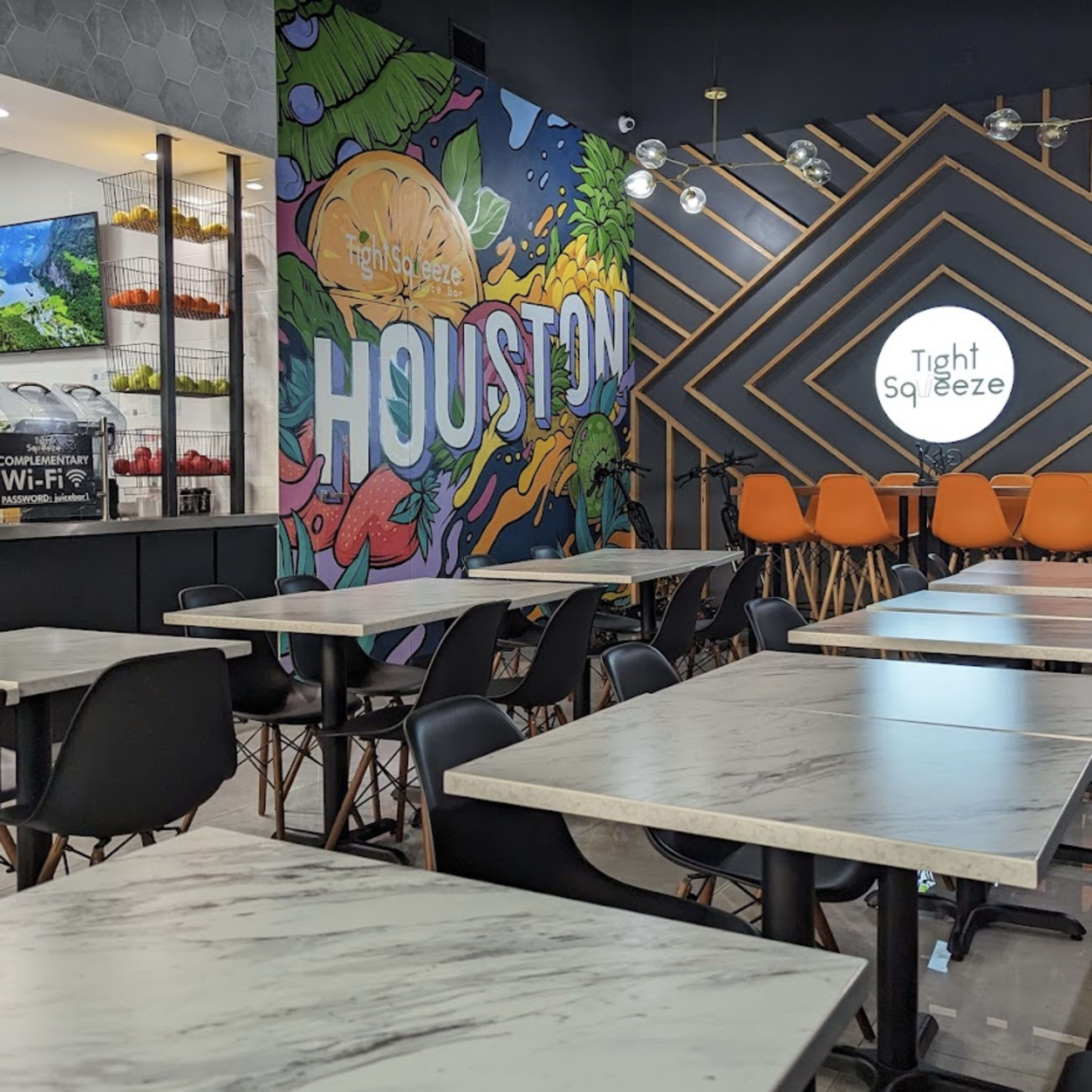A modern café interior with marble-topped tables, black chairs, and a colorful mural featuring the word "HOUSTON" on the wall, alongside orange seating and a service counter.